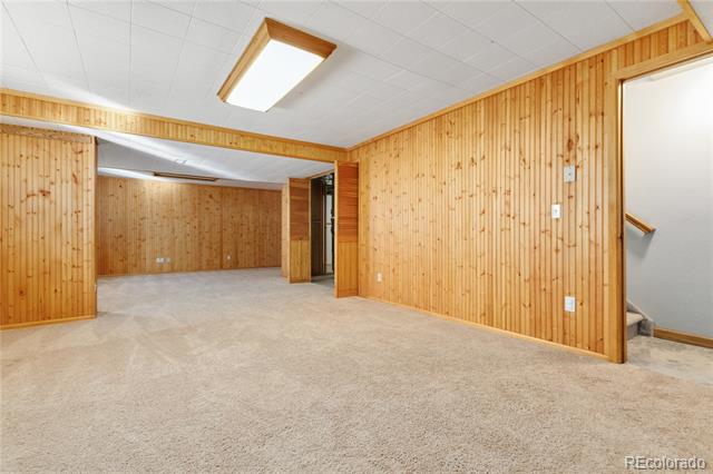 Empty basement room with wood-paneled walls, beige carpet, and an opening to the stairs.