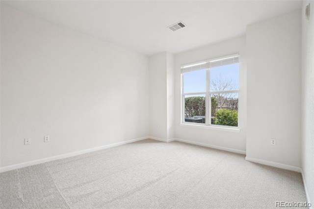 Empty carpeted room with white walls and a large window overlooking shrubs and a parked car.
