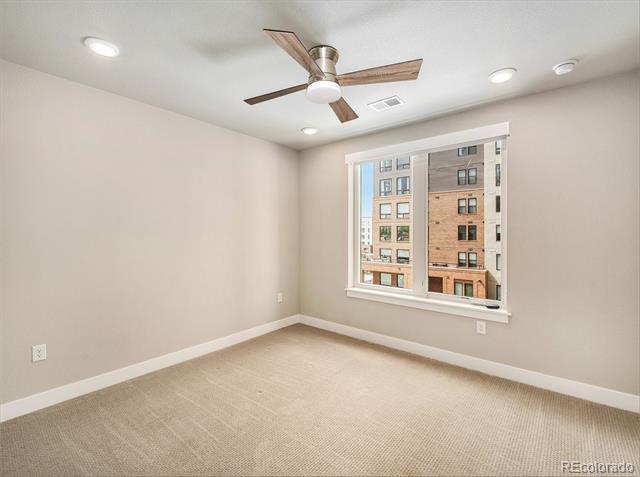 Vacant bedroom with beige carpet, a wood-blade ceiling fan, and a window facing neighboring buildings.