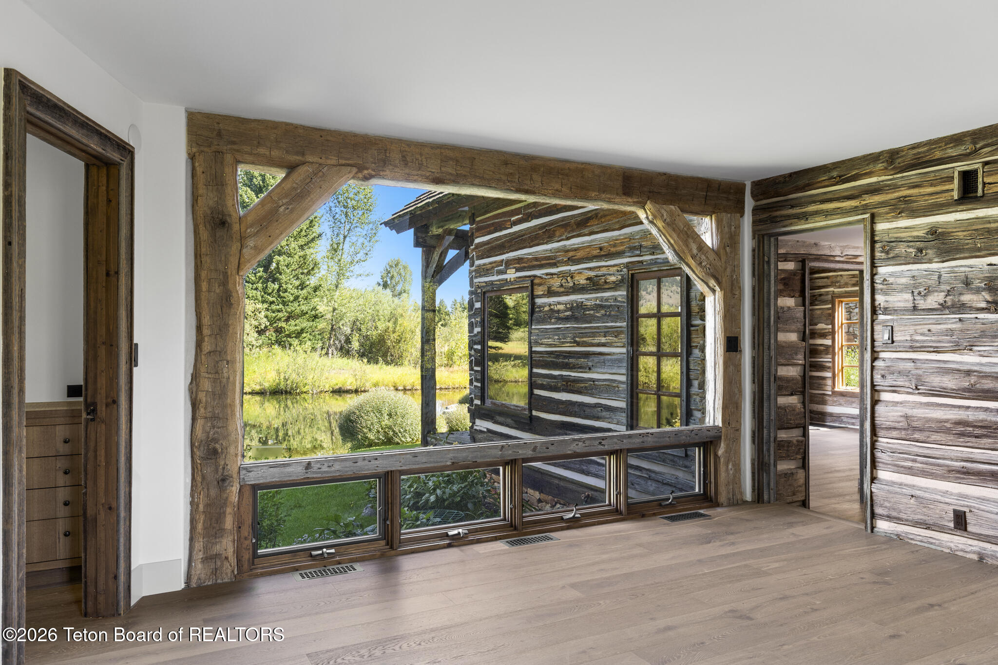 Empty rustic interior with massive timber-framed windows overlooking a pond and log building.