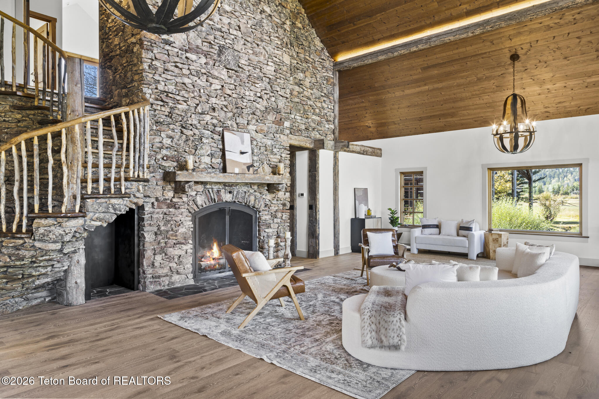 Living room with a towering stone fireplace, rustic staircase, vaulted wood ceiling, and mountain views.