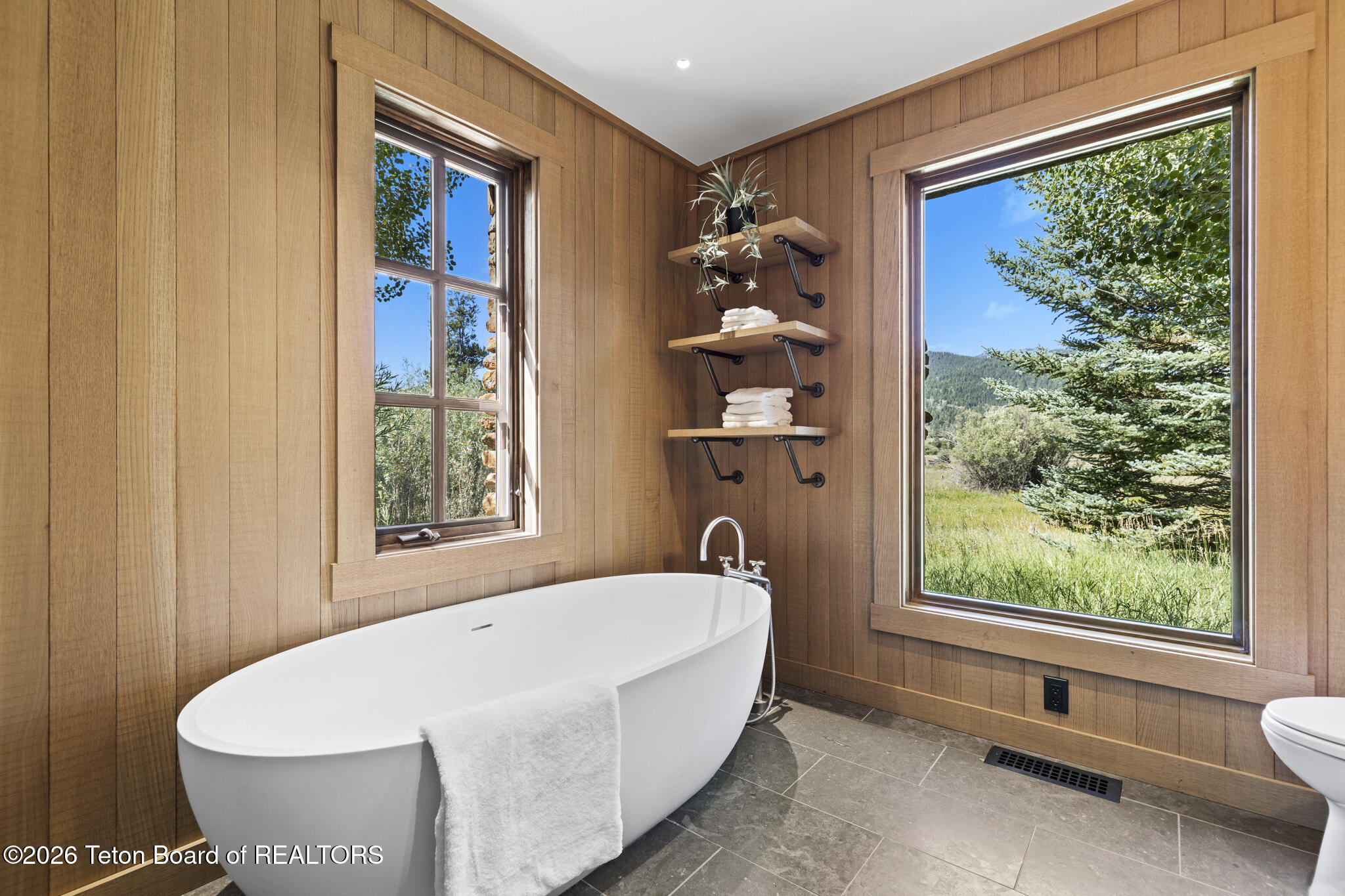 Bathroom with a freestanding soaking tub, wood-paneled walls, and large windows overlooking trees.