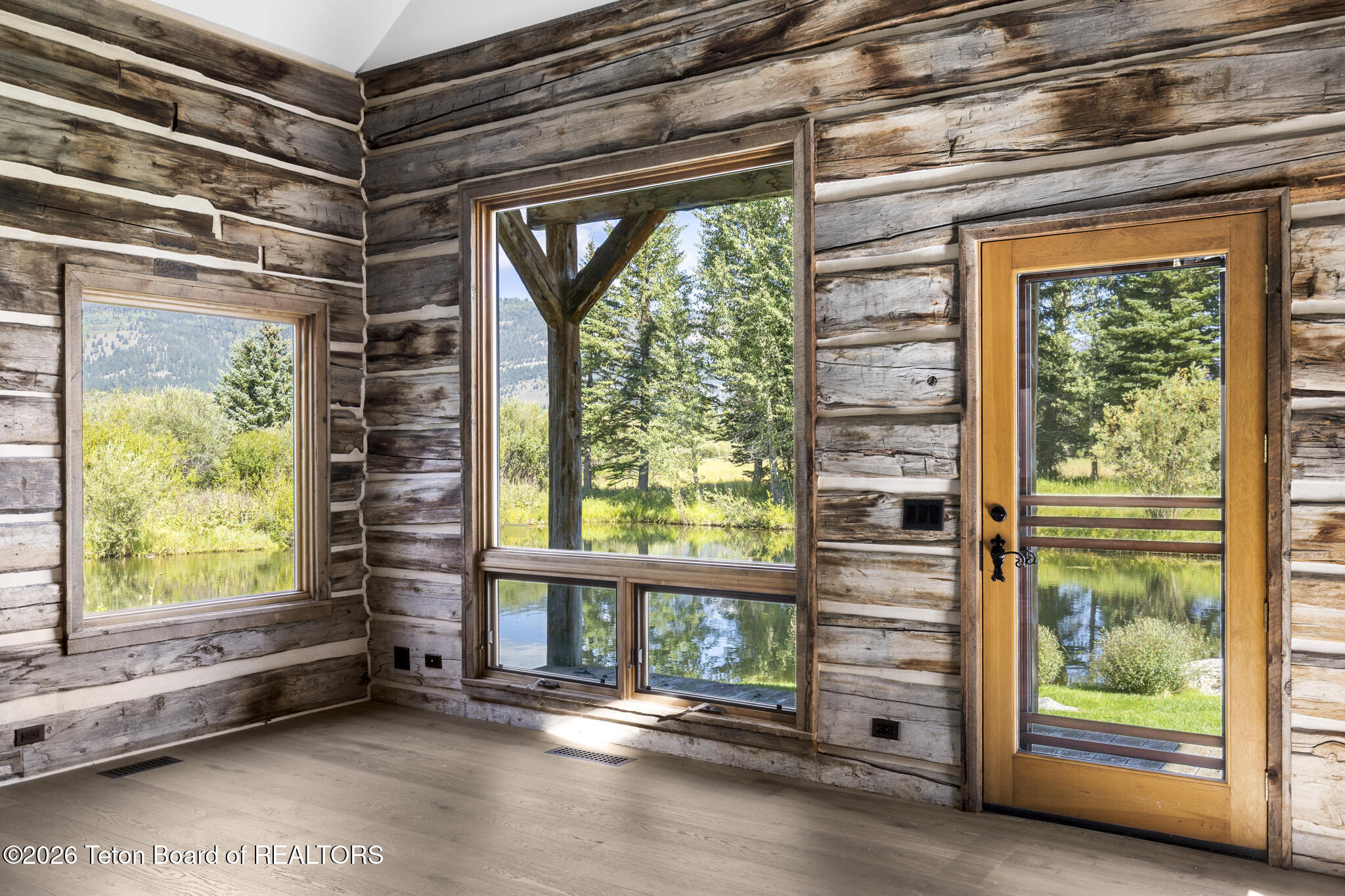 Vacant log cabin room with large windows and a glass door overlooking a pond and trees.