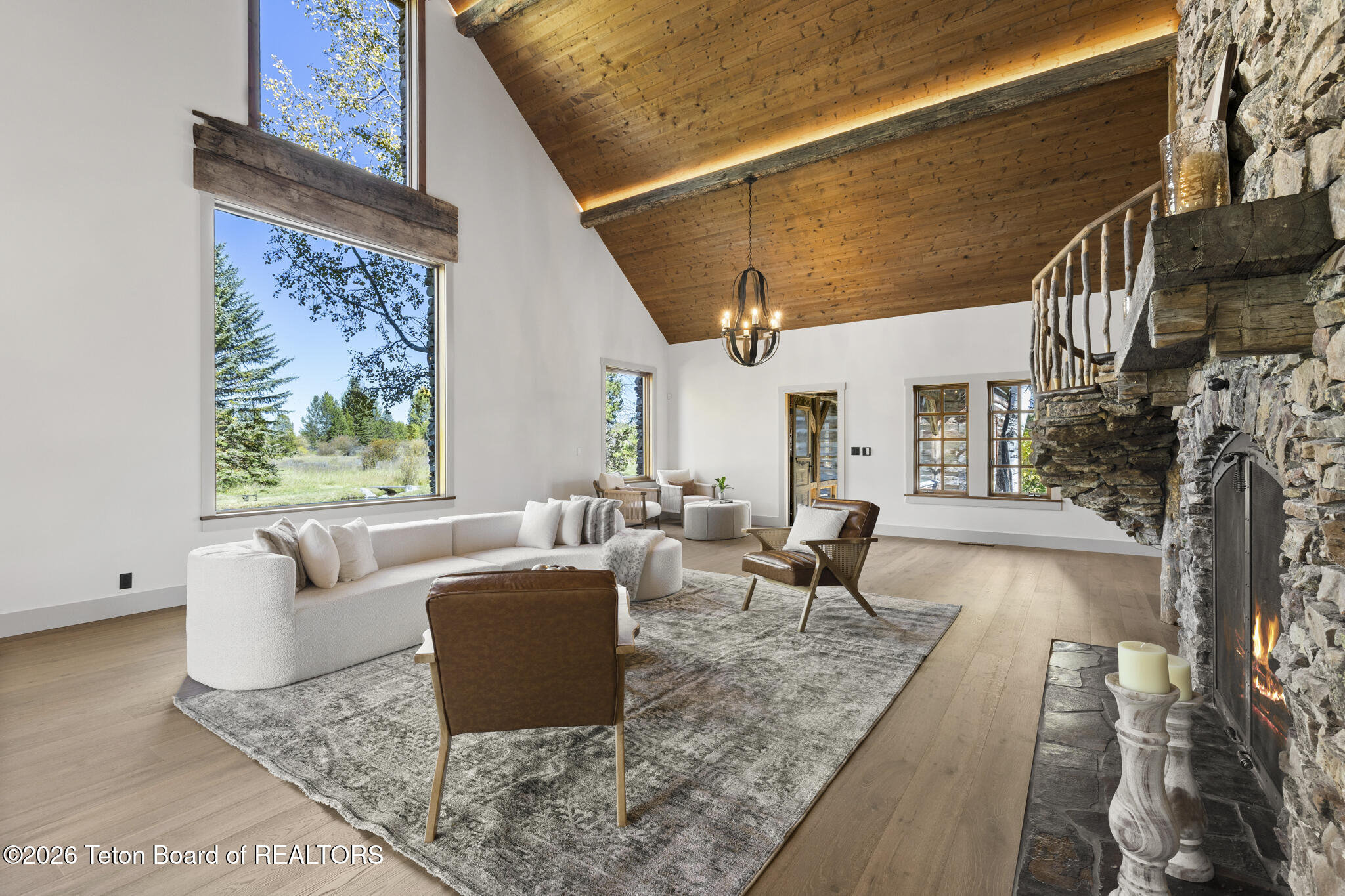 Furnished living room with vaulted wood ceiling, massive stone fireplace, and tall windows overlooking trees.