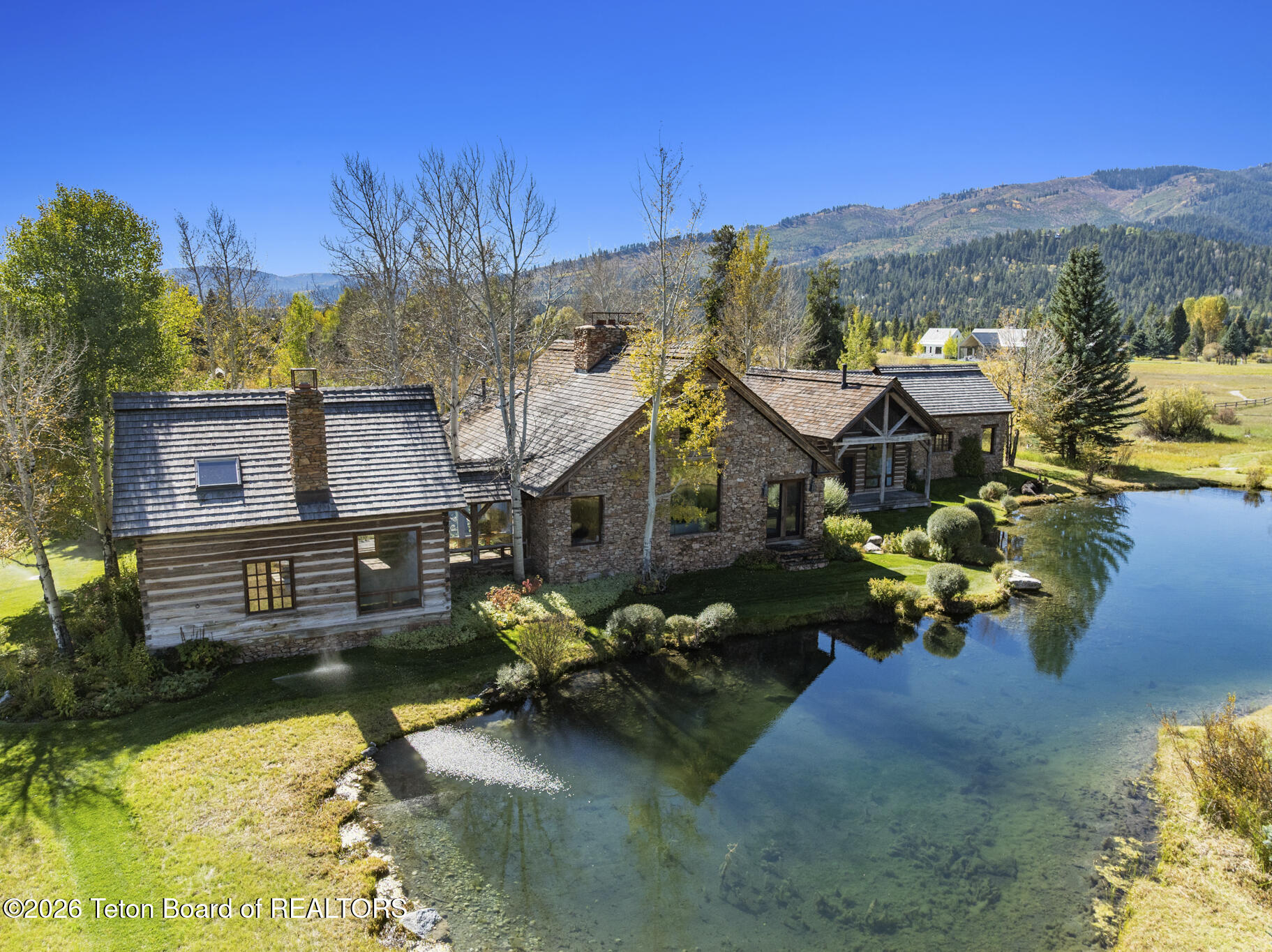 Stone-and-timber home beside a clear pond, with landscaped grounds and forested mountains beyond.