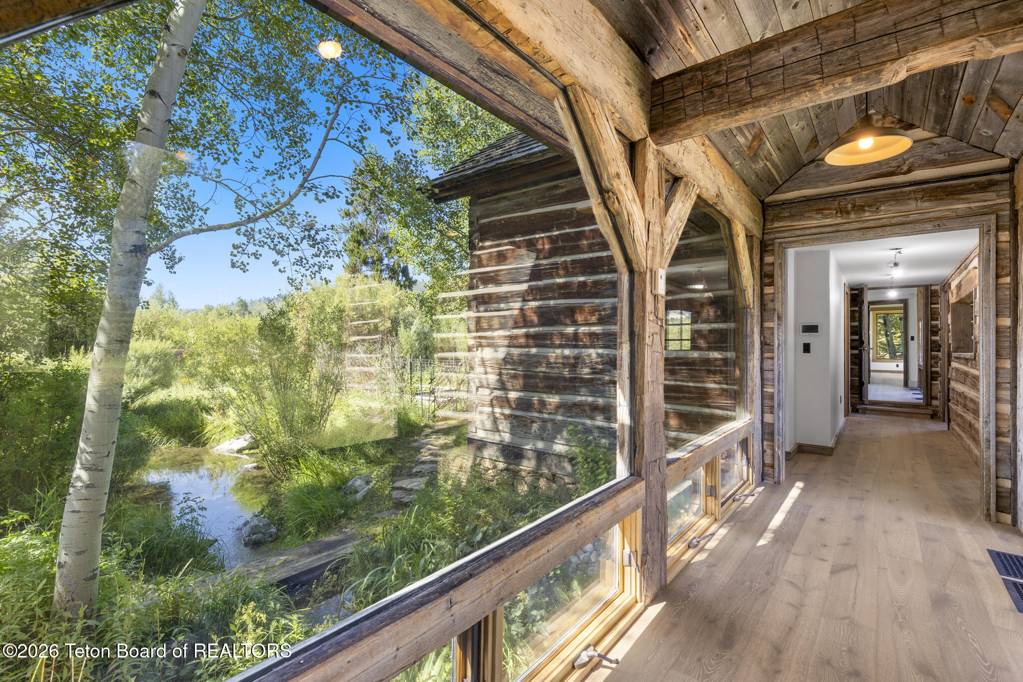 Wood-framed hallway with expansive windows overlooking a garden stream and trees.