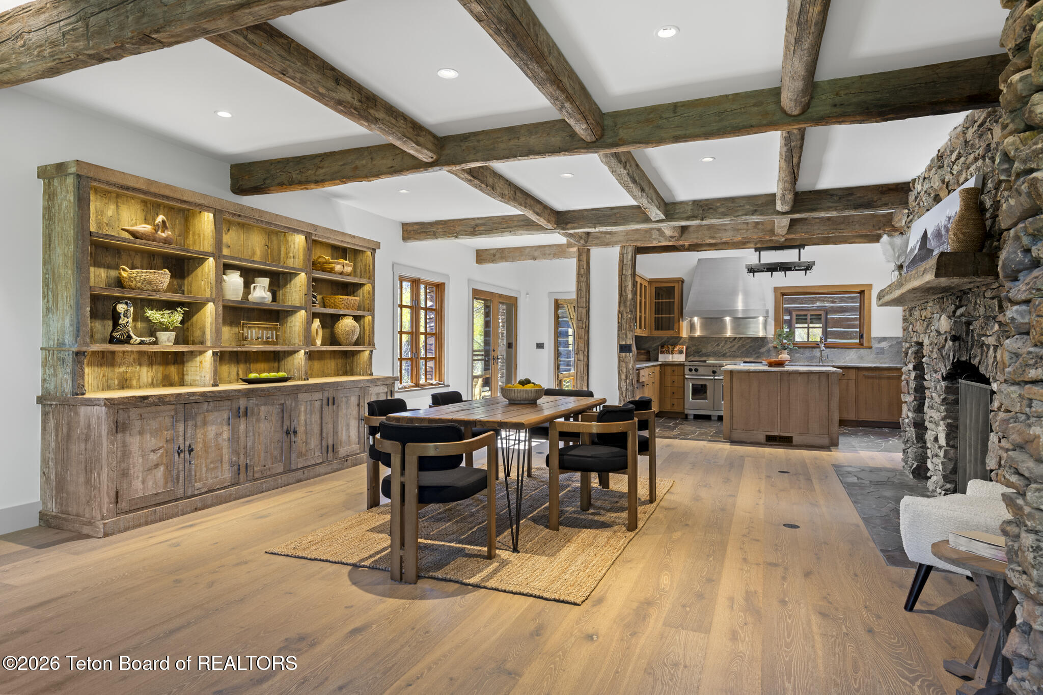 Open-plan kitchen and dining room with exposed timber beams, rustic shelving, and a stone fireplace.