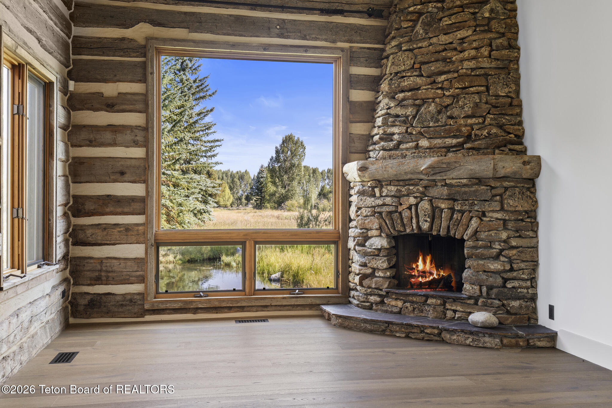 Log home interior with a stone fireplace beside a large window overlooking a meadow and pond.