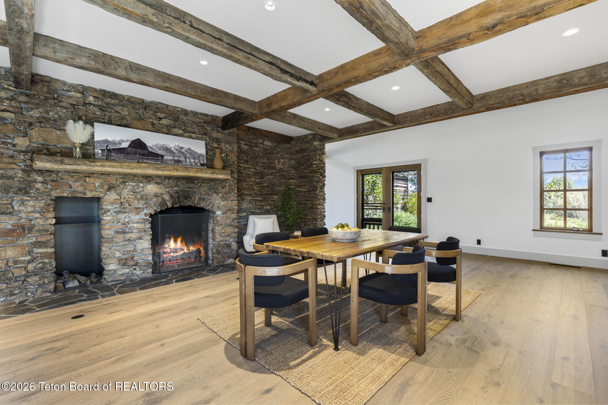Dining room with exposed timber beams, a stone fireplace, and a wooden table beside large windows.