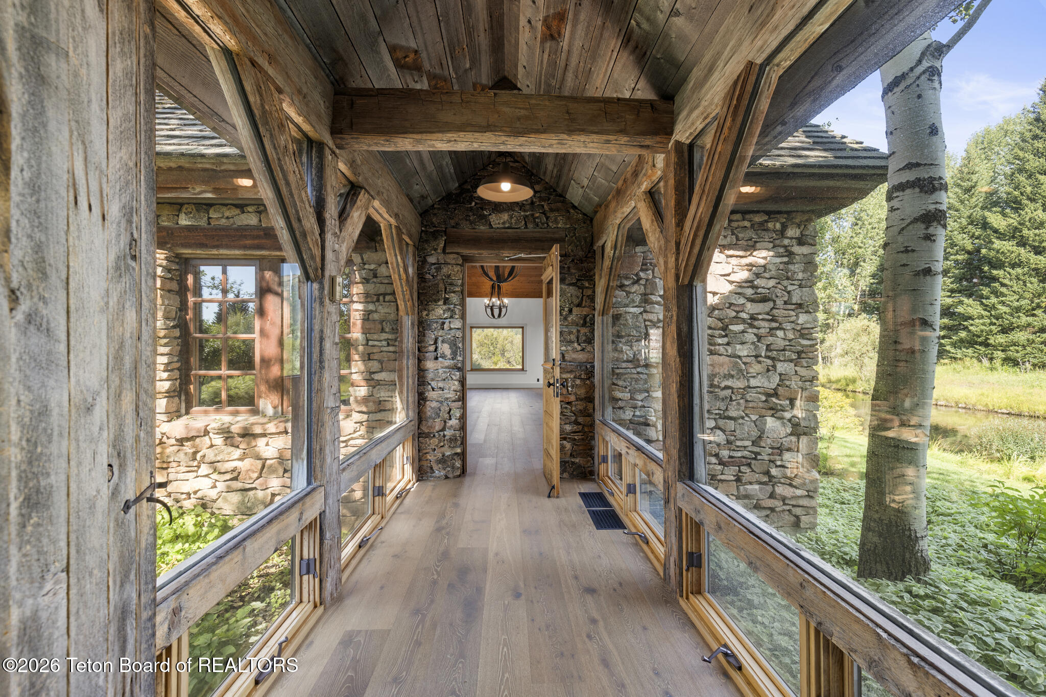 Enclosed breezeway with vaulted timber beams, stone walls, and large windows overlooking leafy grounds.