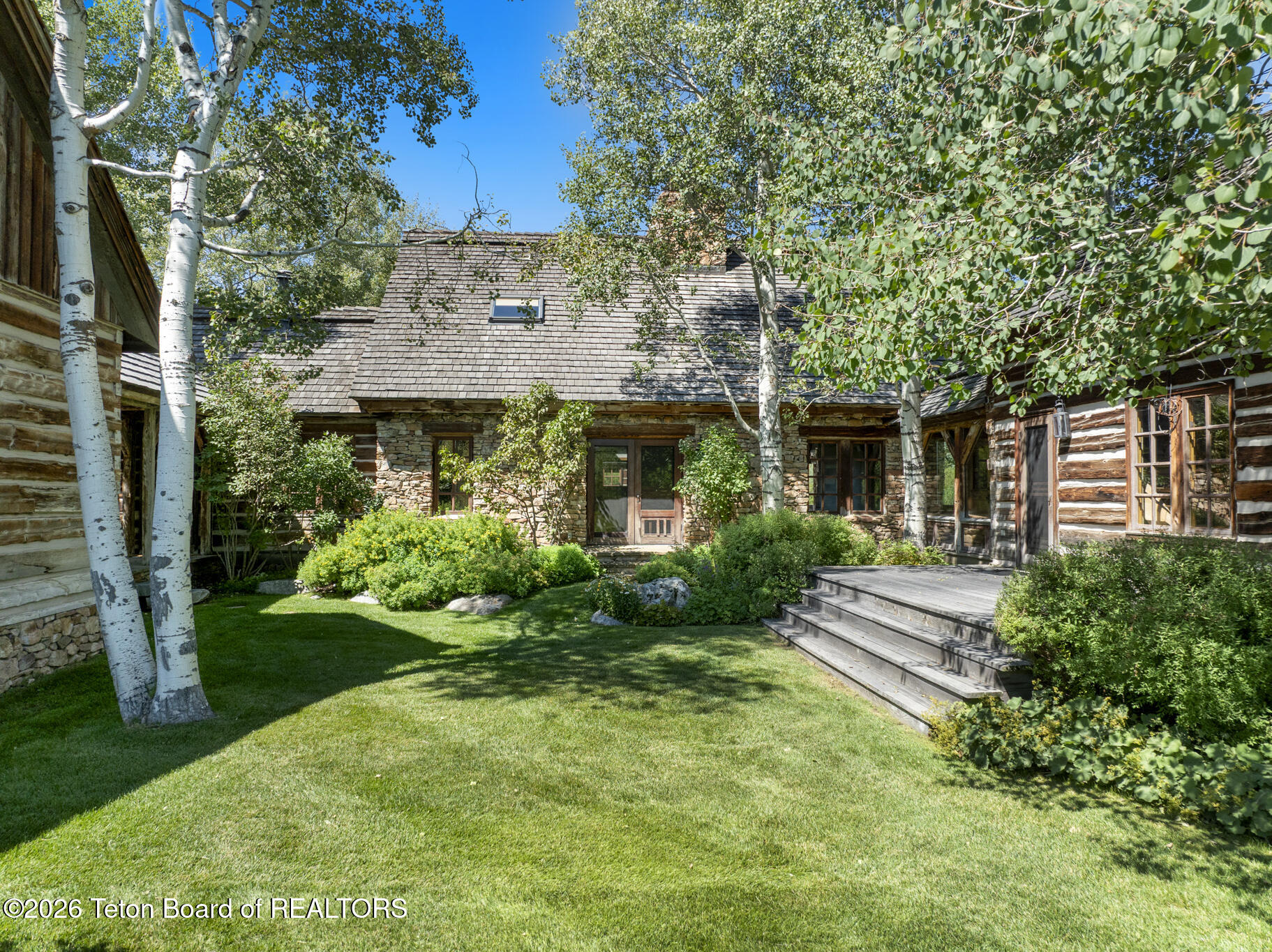 Rustic log-and-stone house framed by aspens, with a green lawn and raised wooden deck.