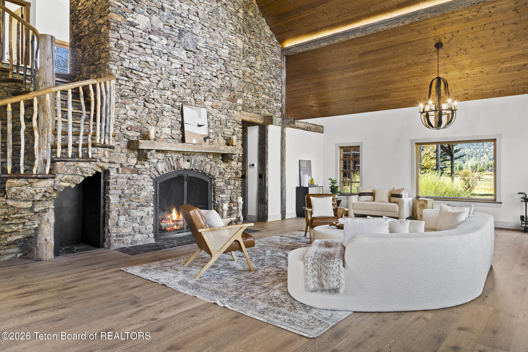 Living room with a towering stone fireplace, vaulted wood ceiling, rustic loft staircase, and curved white sofa.