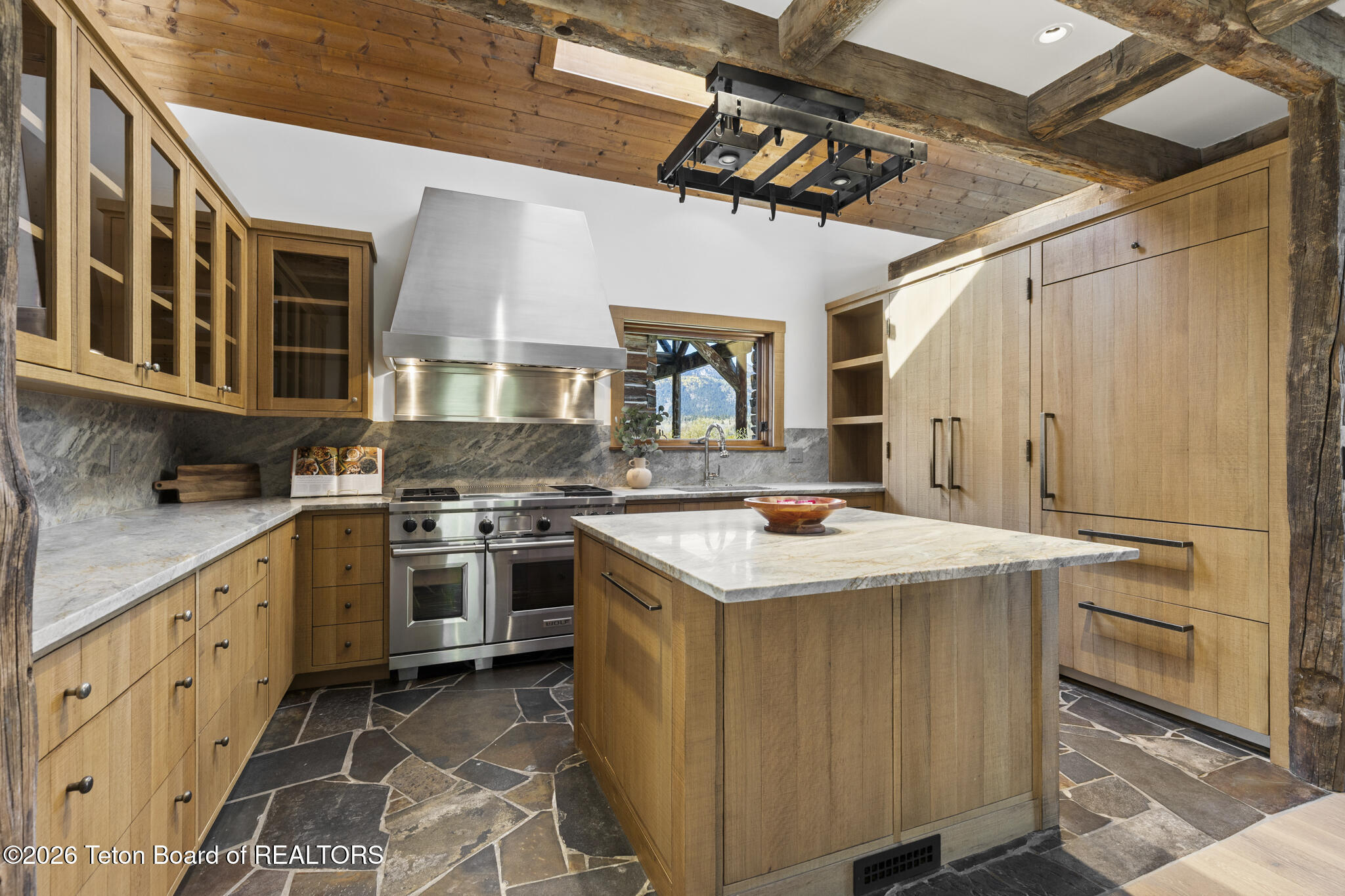 Kitchen with a stone-topped island, light wood cabinets, slate floors, and exposed timber ceiling beams.