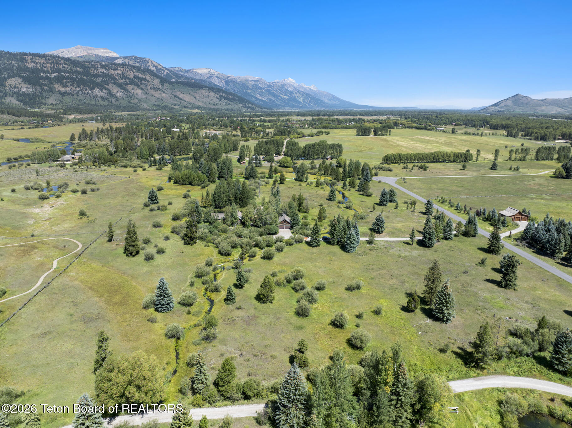Aerial view of a rural property with open meadows, a winding creek, scattered homes, and mountains.