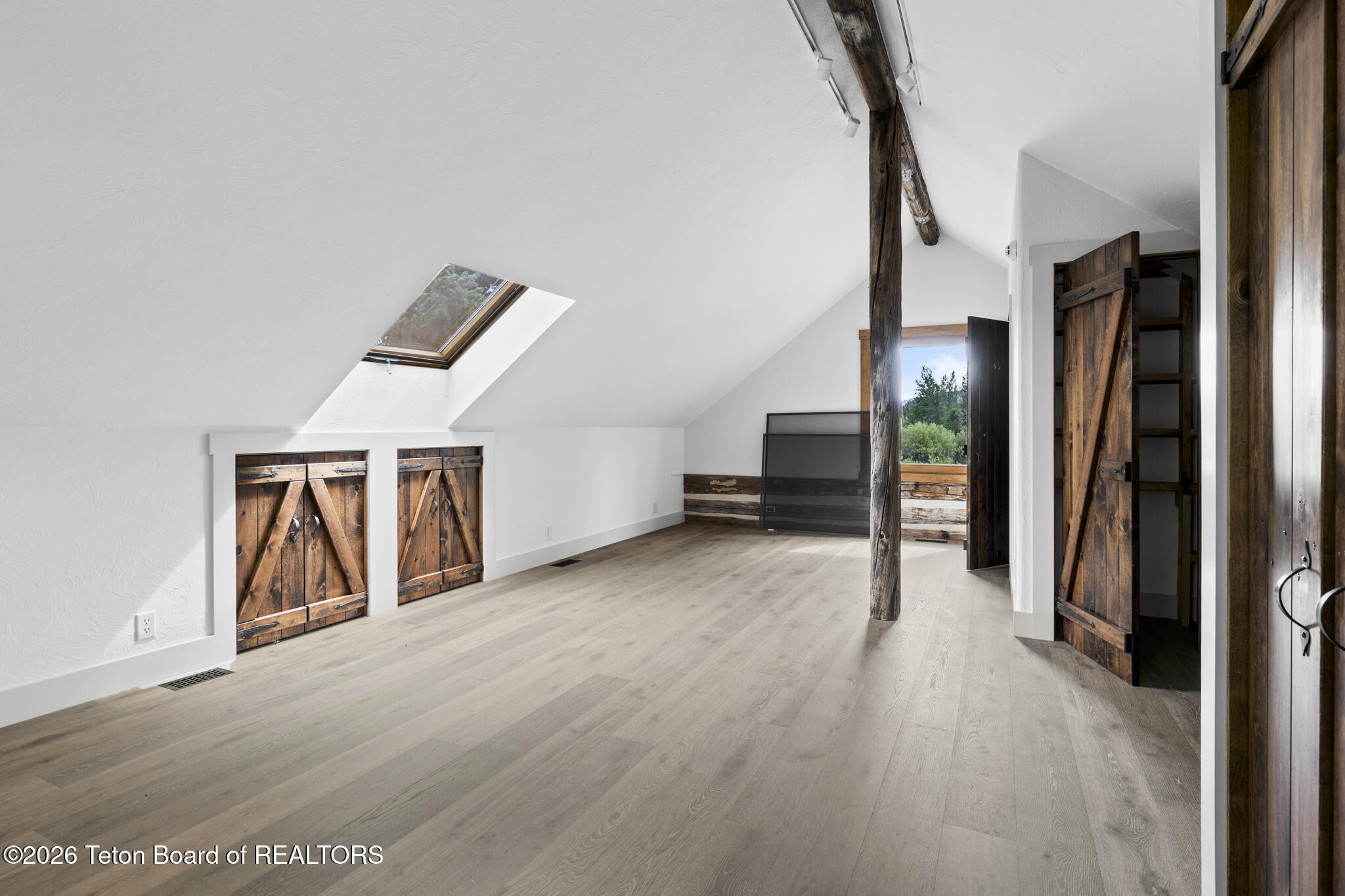 Empty loft with pale wood floors, a skylight, exposed timber beams, and rustic barn doors.