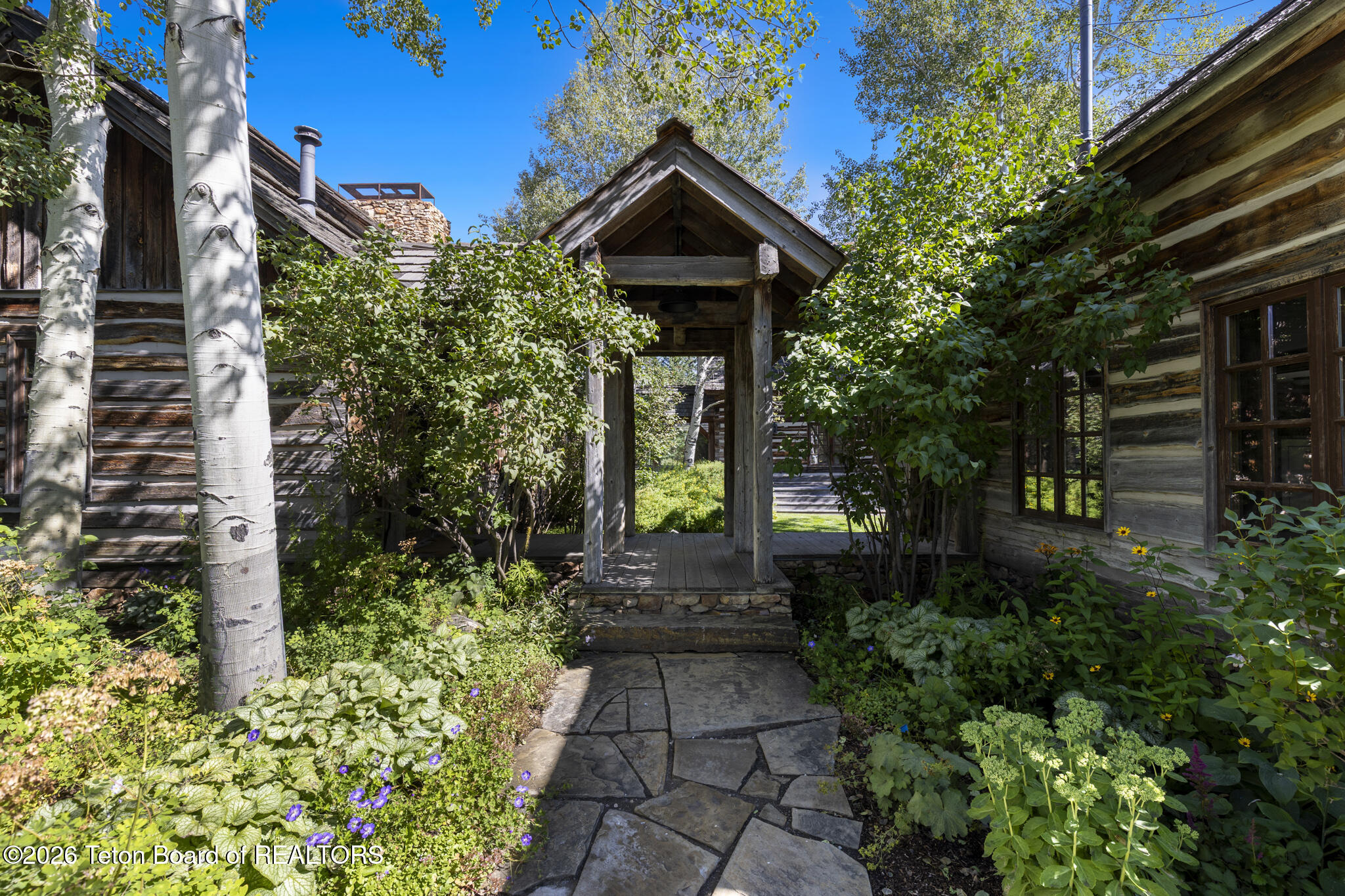 Rustic log home entrance framed by aspens and gardens, with a timber entryway and stone walkway.