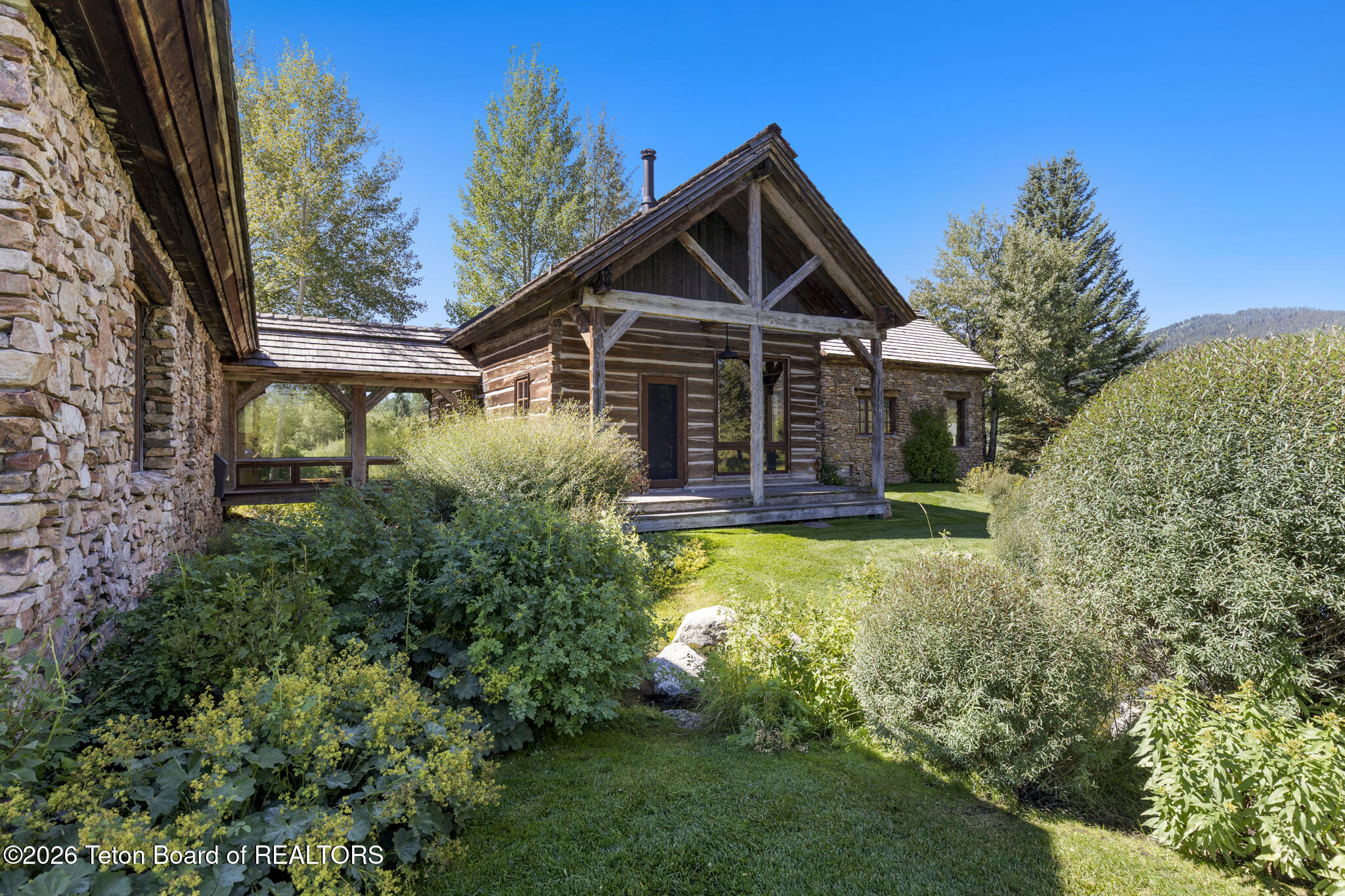 Rustic log-and-stone home with a timber porch, landscaped garden, and wooded mountain backdrop.