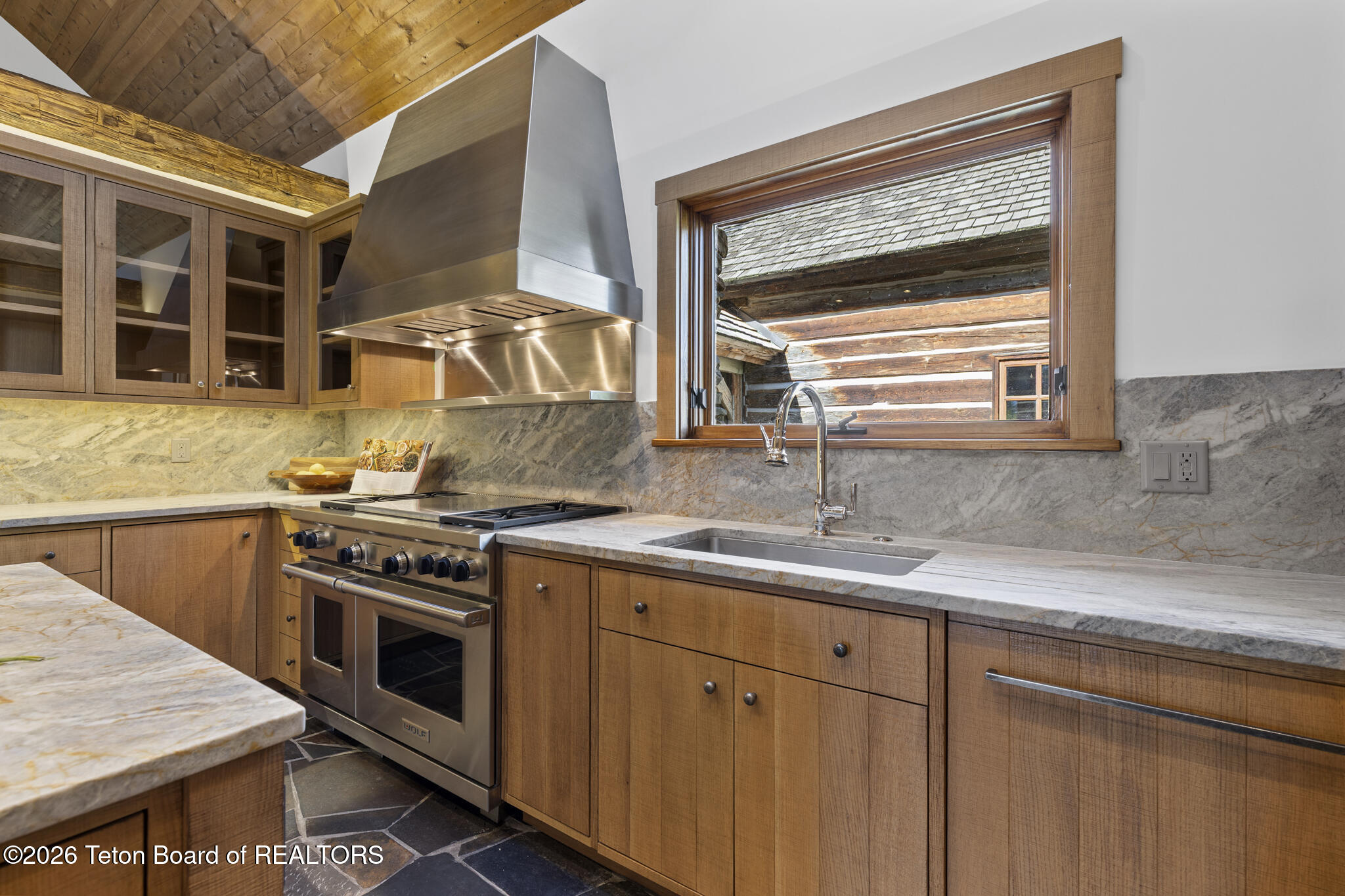 Kitchen with warm wood cabinetry, marble counters, stainless steel range, and a window overlooking a log building.