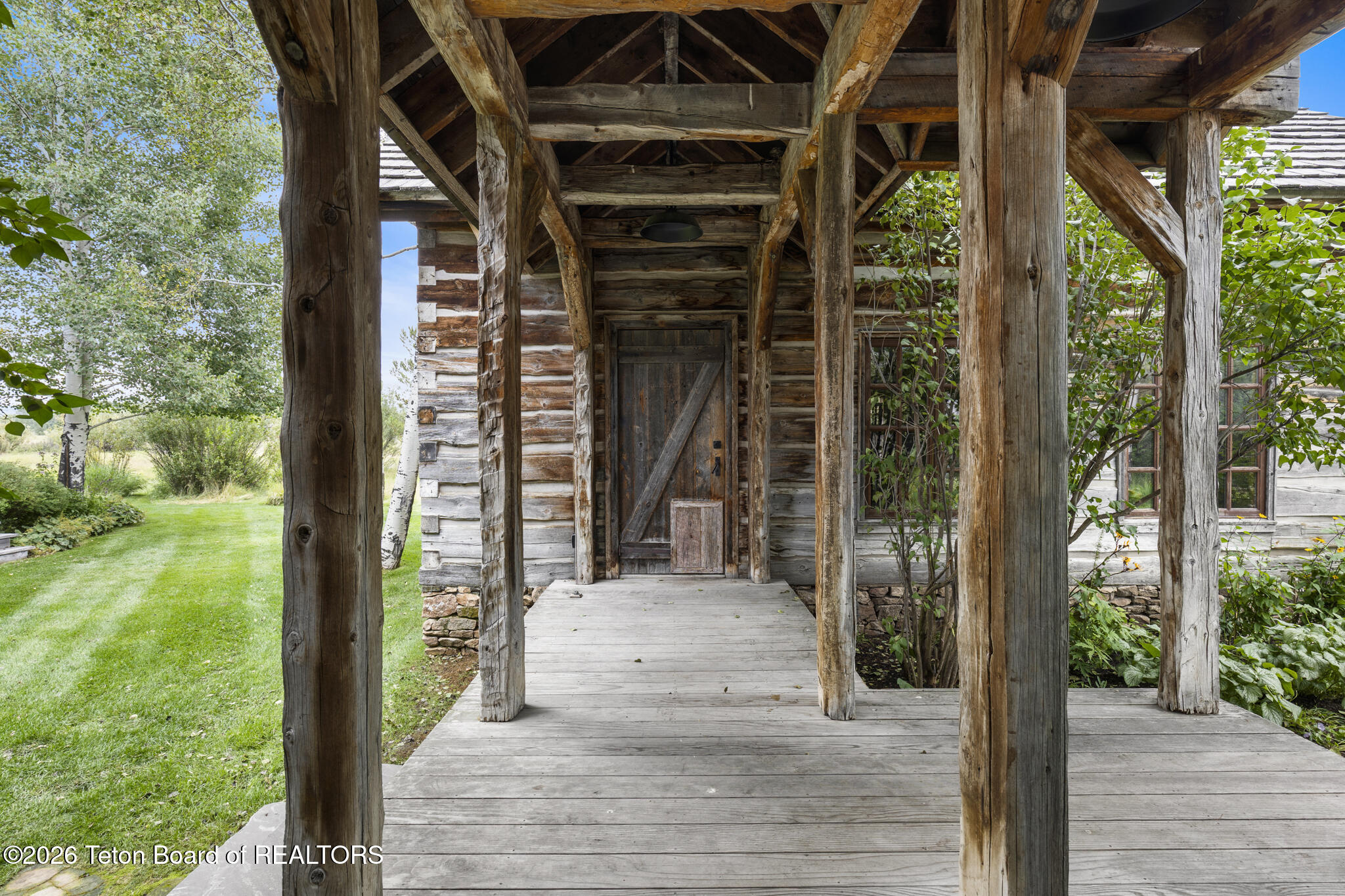 Rustic log home entrance beneath a timber-framed porch, with a weathered wooden door and green garden.