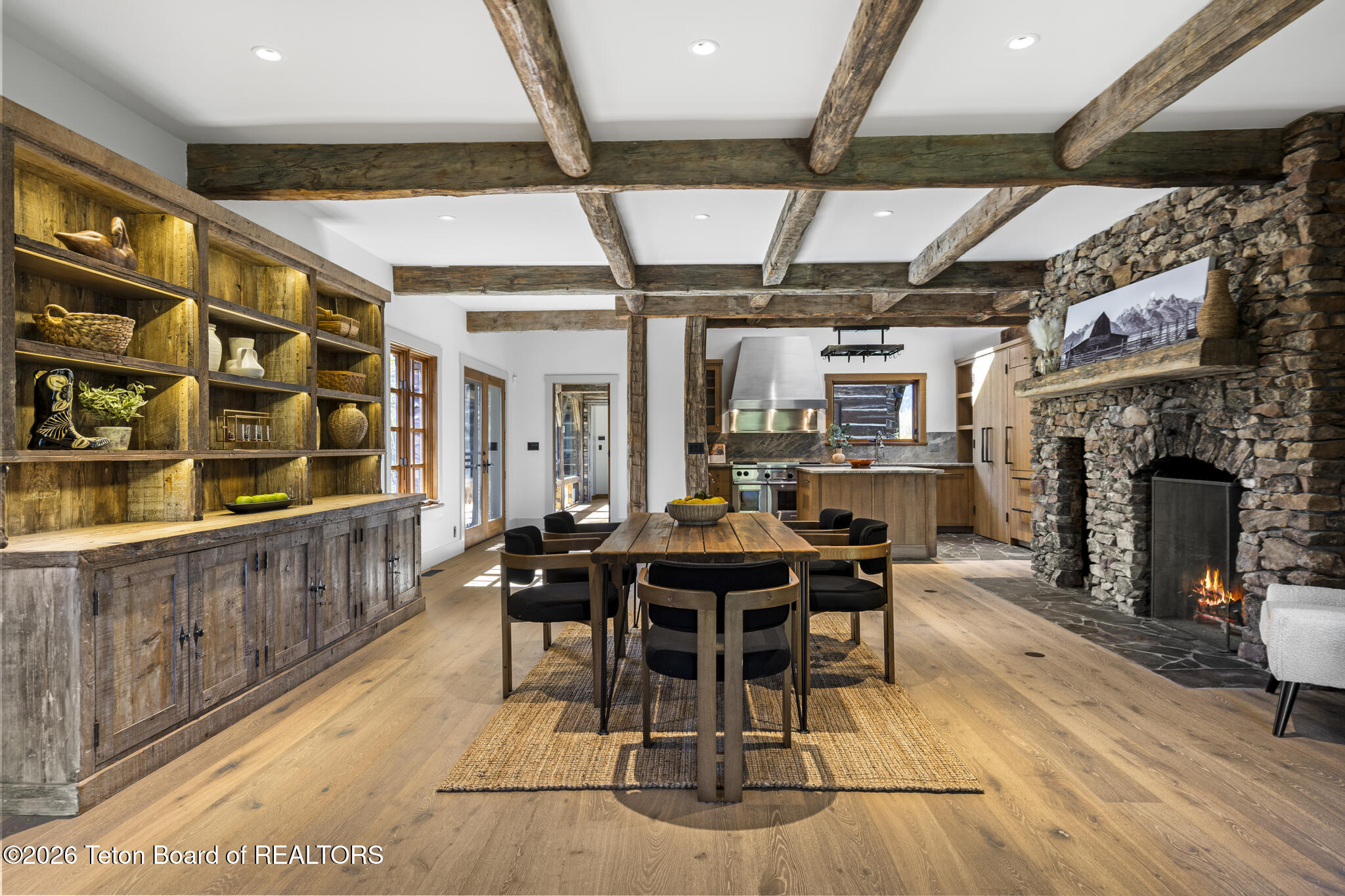 Open-plan kitchen and dining room with exposed timber beams, built-in shelving, and a stone fireplace.