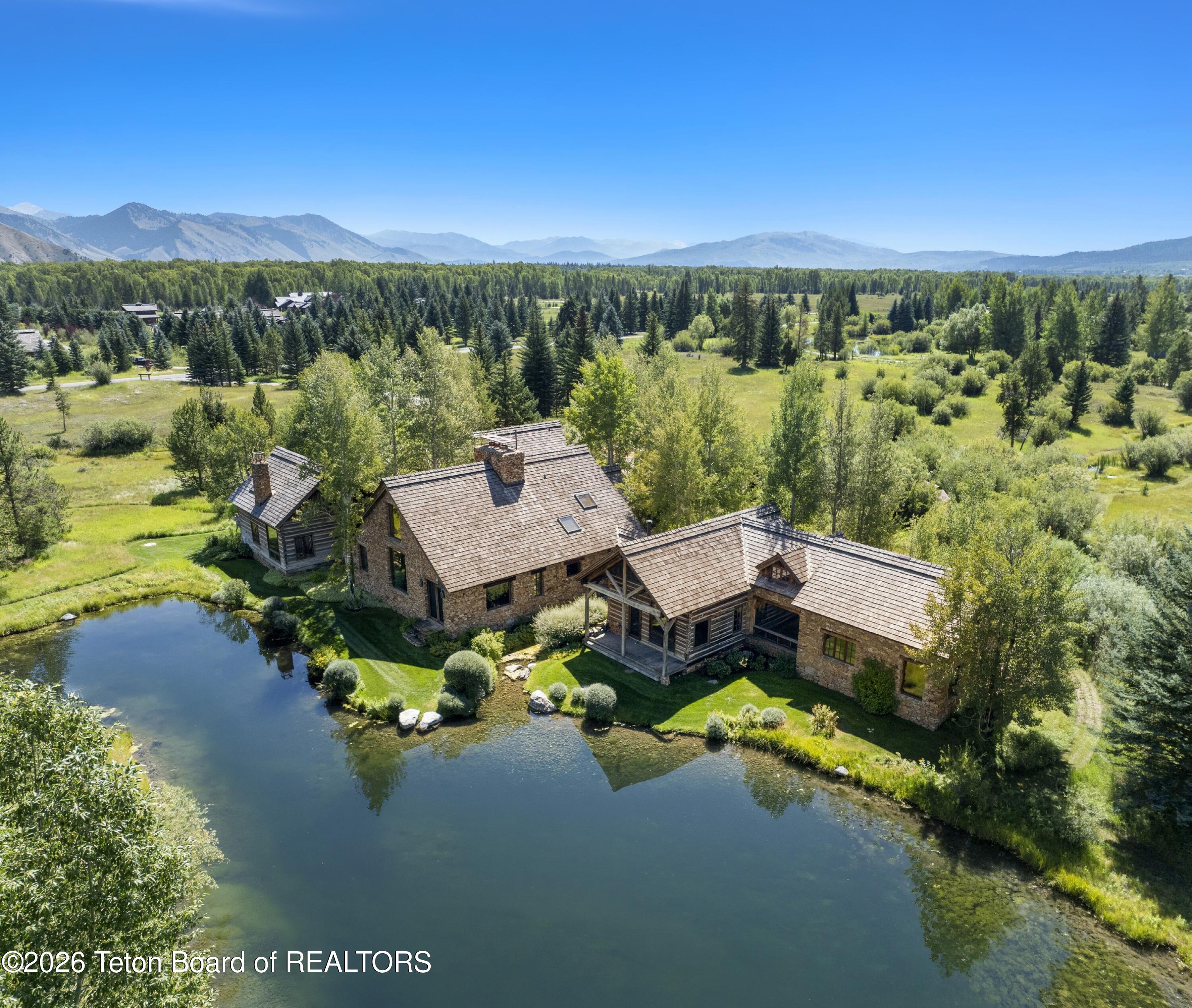 Aerial view of a stone-and-timber home beside a pond, with forest and mountains beyond.
