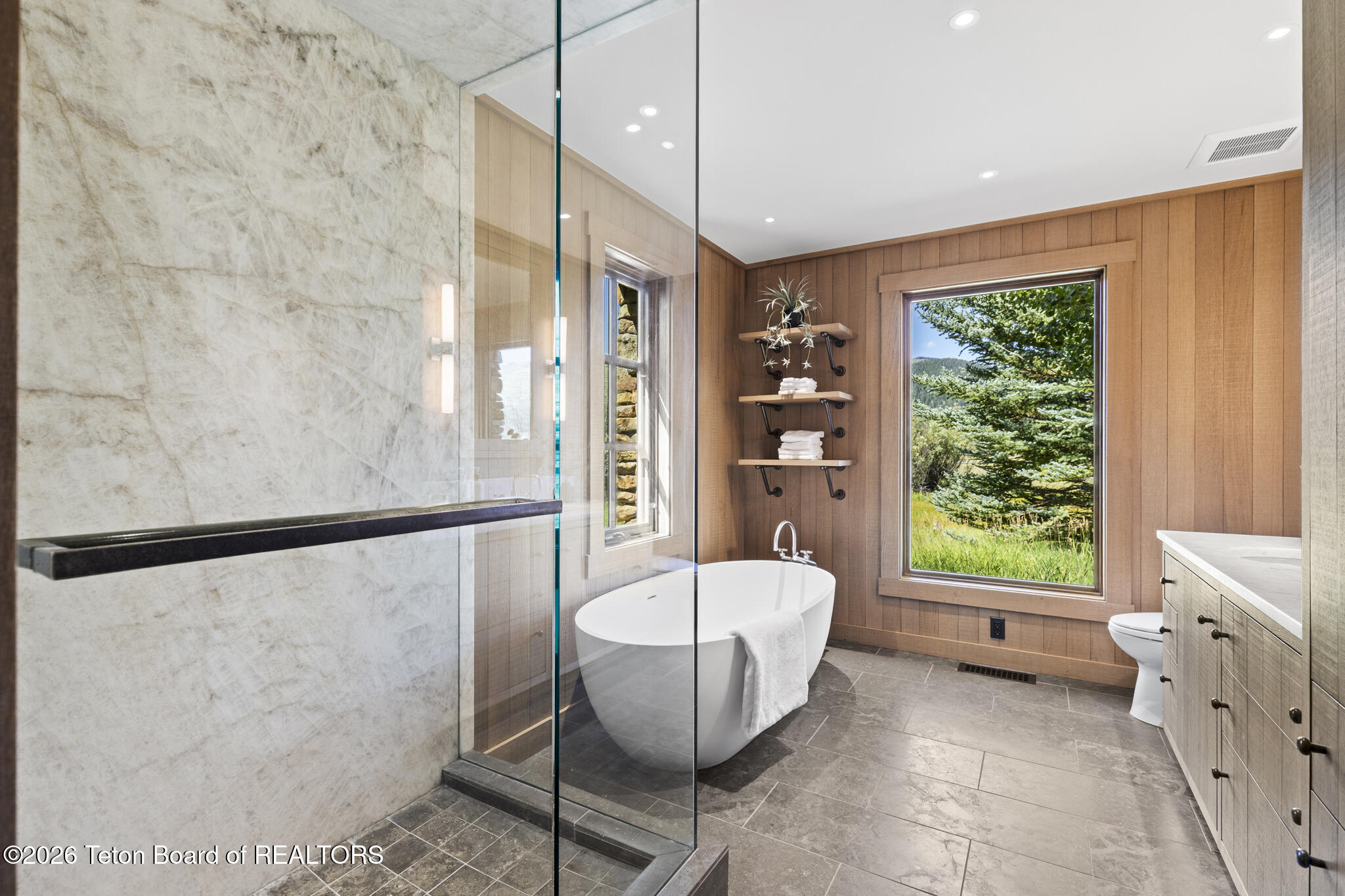 Bathroom with a glass shower, freestanding tub, wood-paneled walls, and a large window overlooking trees.