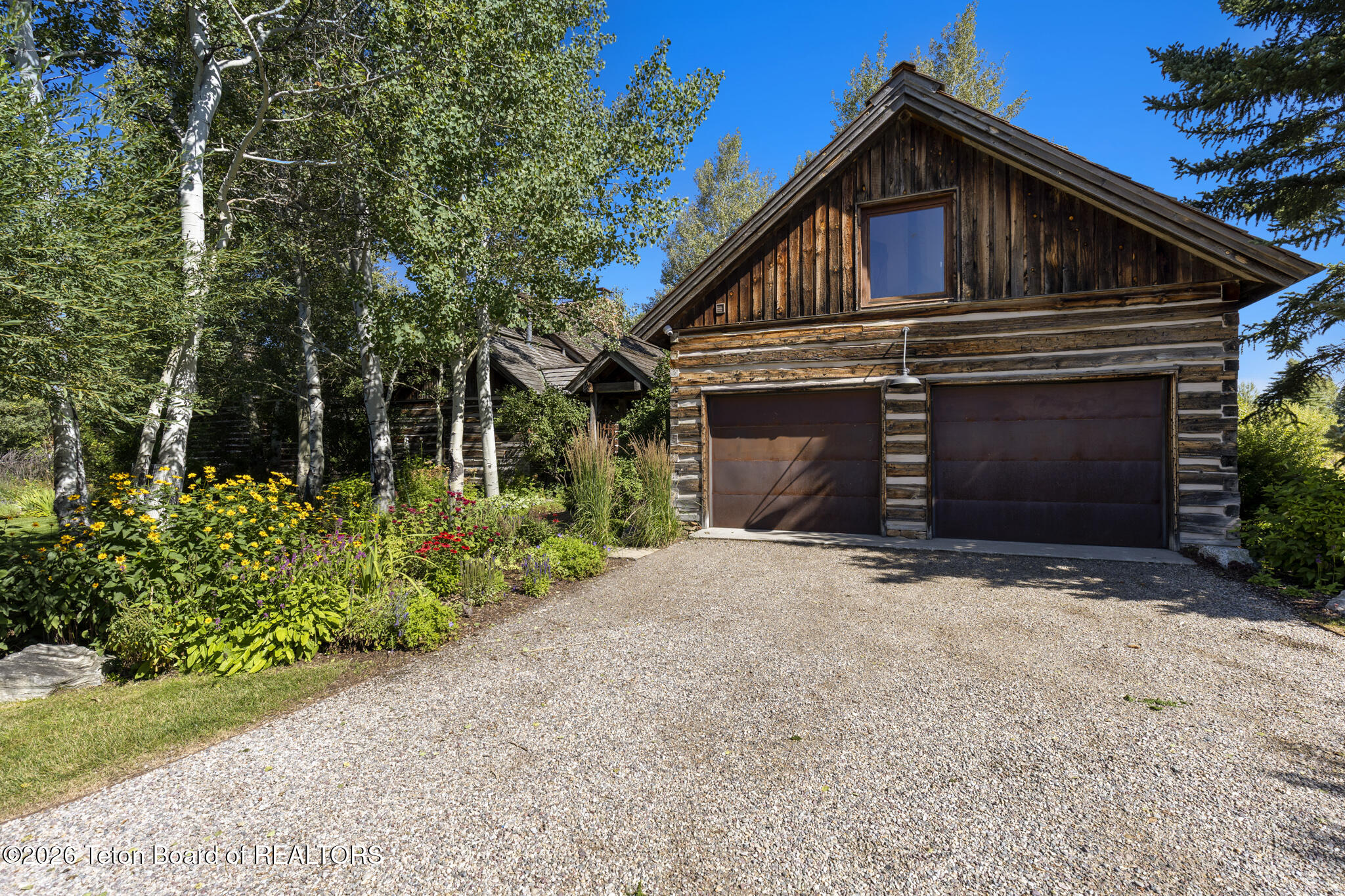 Rustic timber garage with two doors, a gravel driveway, and colorful flower beds beneath mature trees.