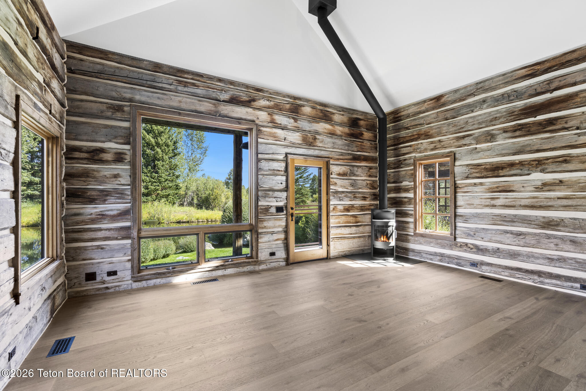 Vacant log cabin living room with broad windows, pale wood floors, and a lit freestanding stove.