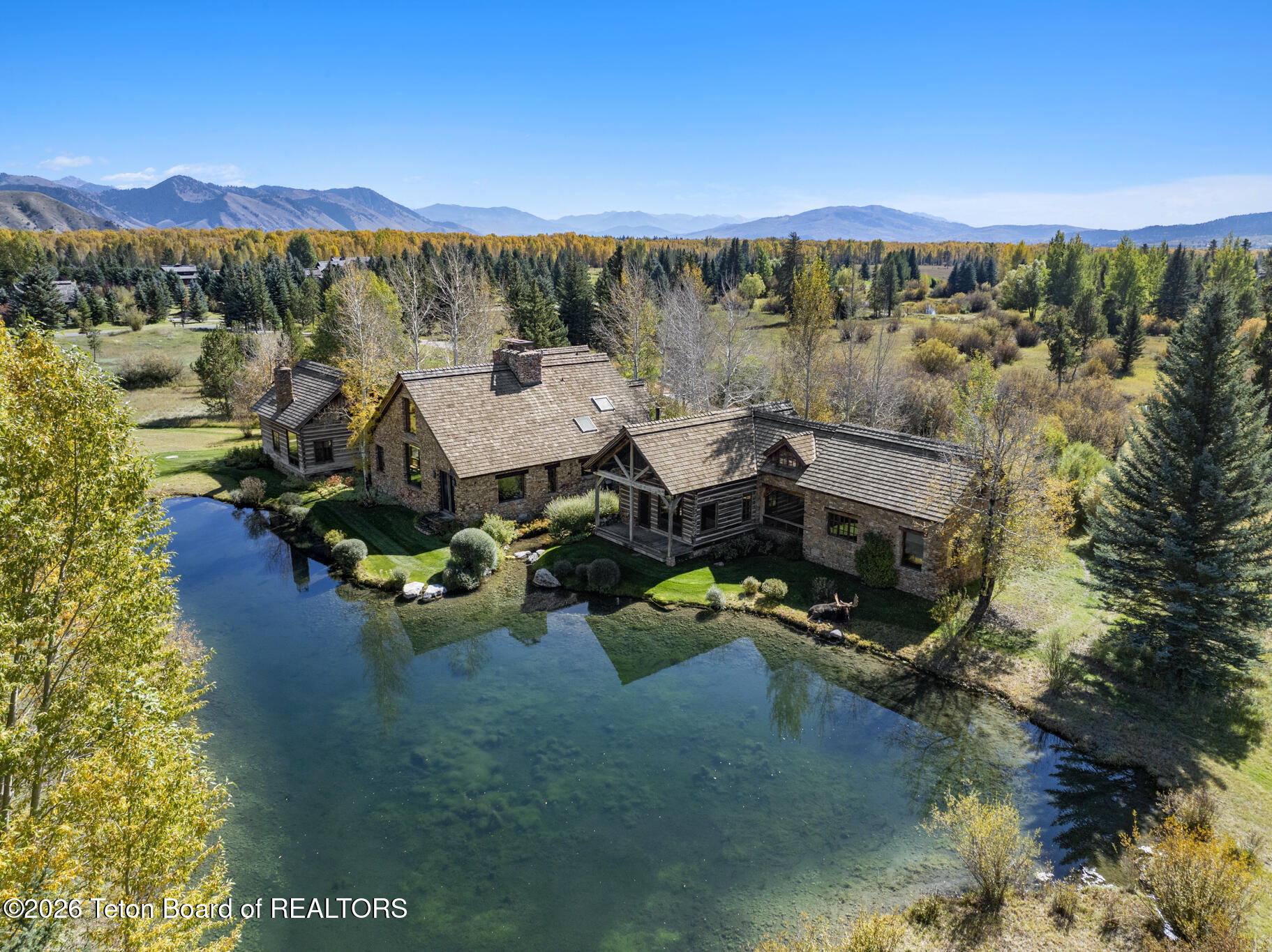 Aerial view of a stone-and-timber home beside a pond, with autumn trees and mountain views.