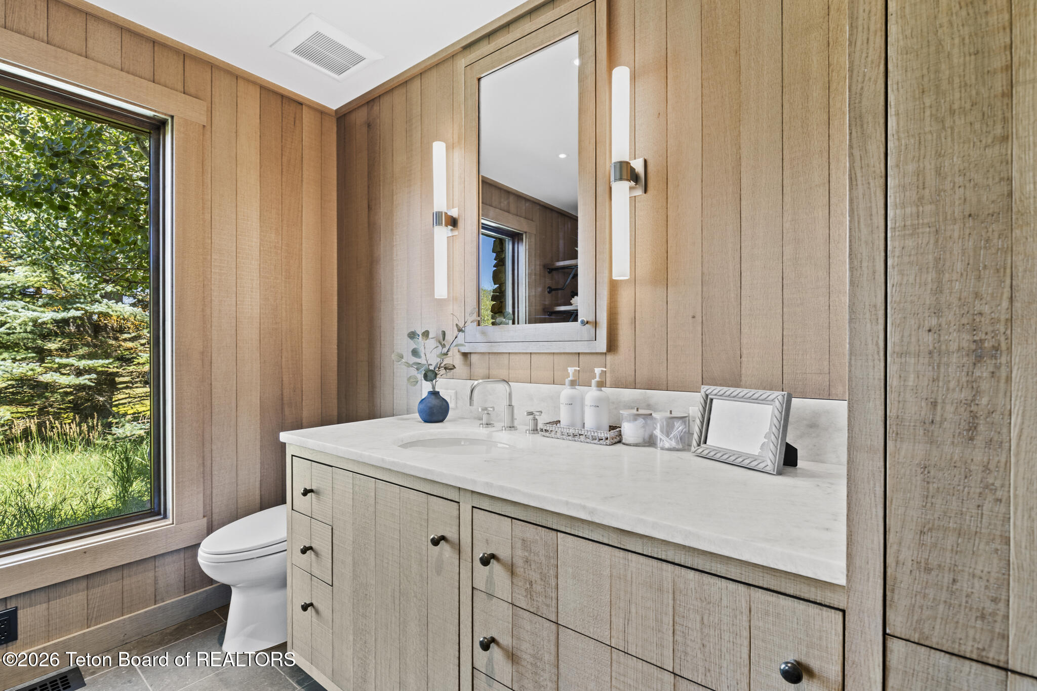Bathroom with a light wood vanity, white countertop, and large window overlooking trees.