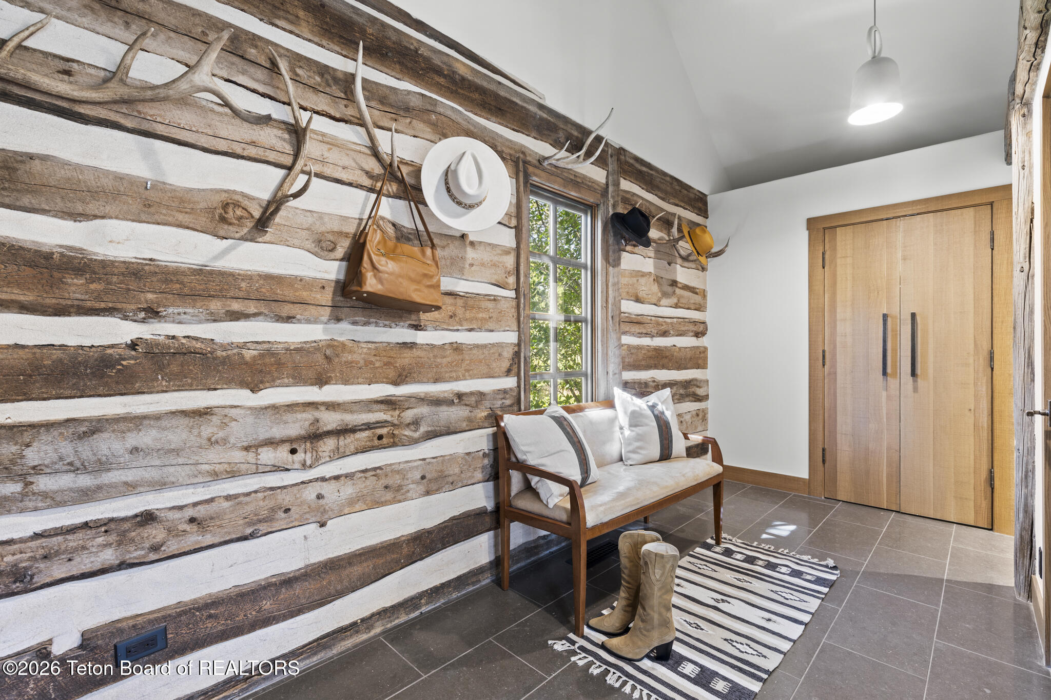 Entryway with a whitewashed log wall, antler hooks, a wood bench, boots, and tall wooden closet doors.