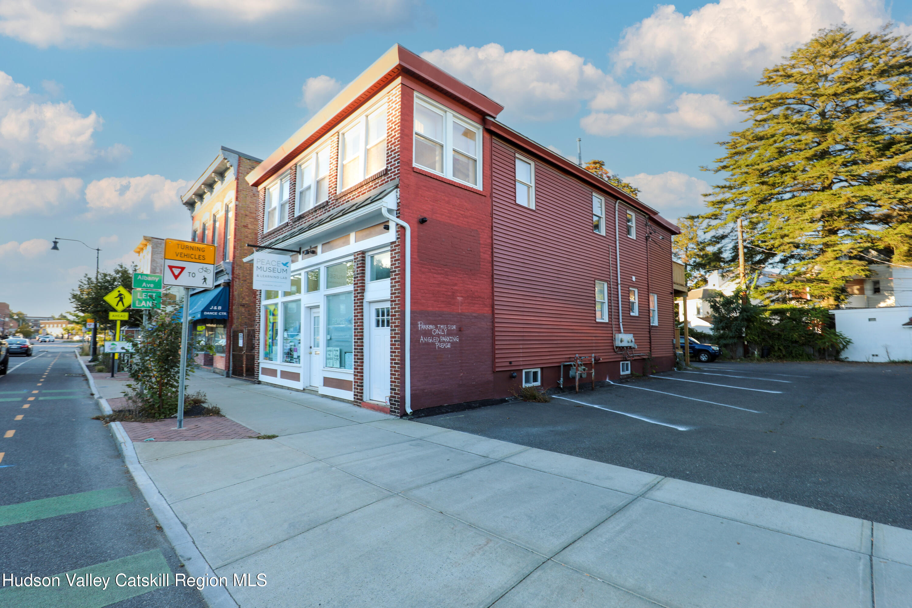 Two-story red corner building with a white storefront, sidewalk, and adjacent parking lot.