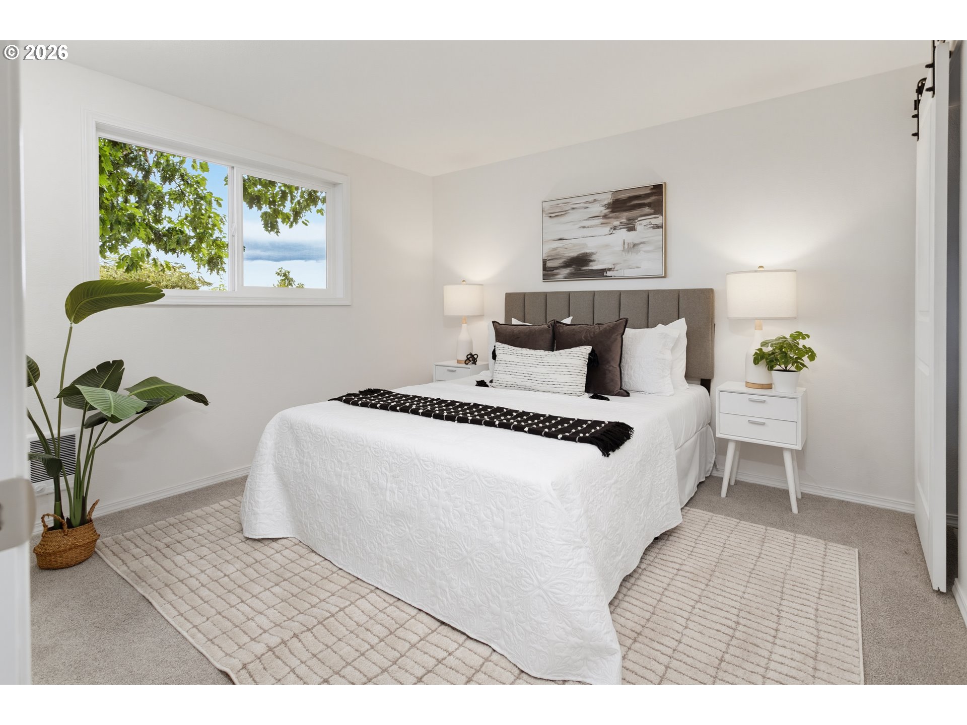 Staged bedroom with a white bed, gray headboard, leafy window view, and light grid-pattern rug.