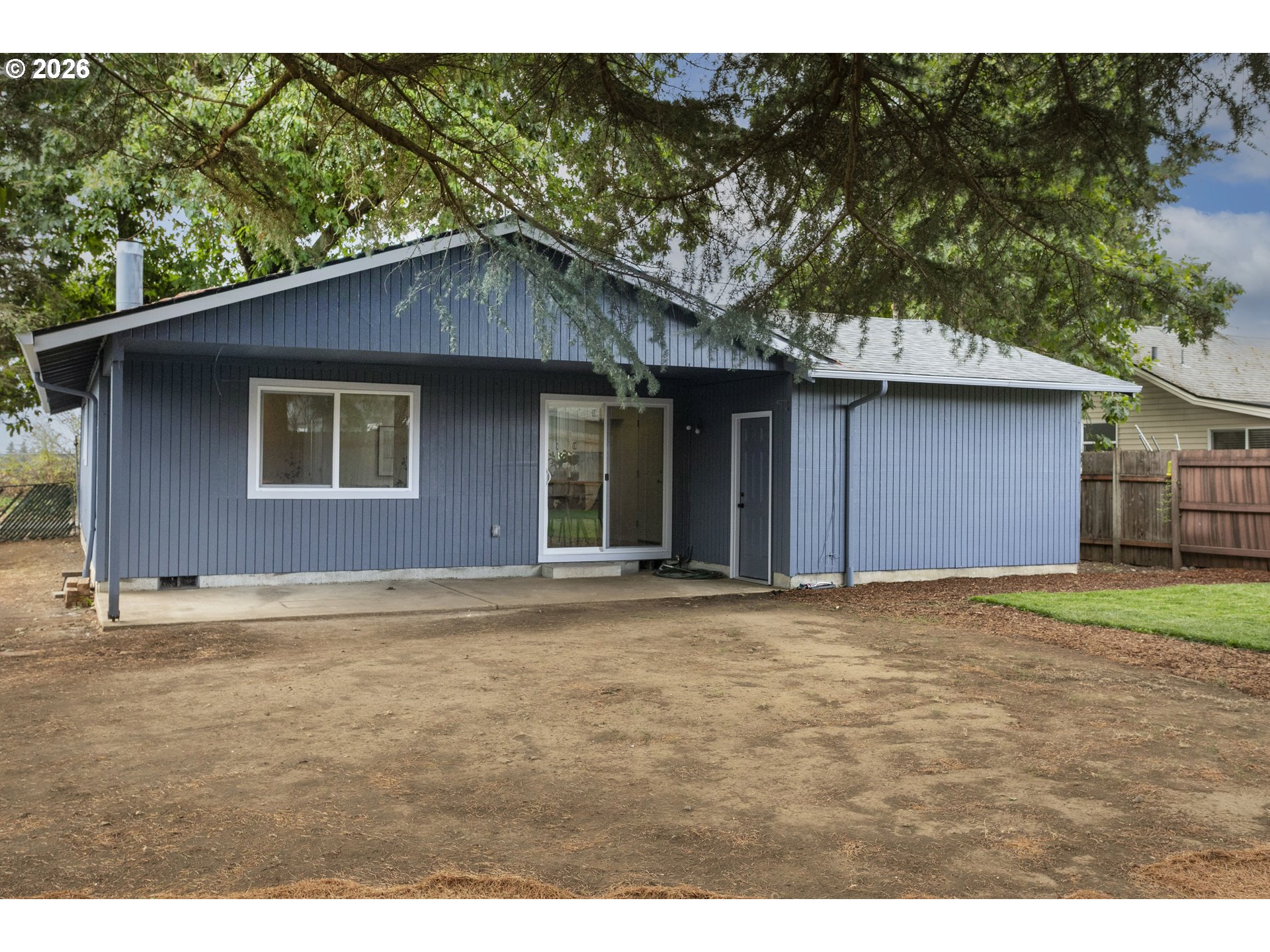 Blue-sided house with a covered patio and sliding glass door beneath mature trees.