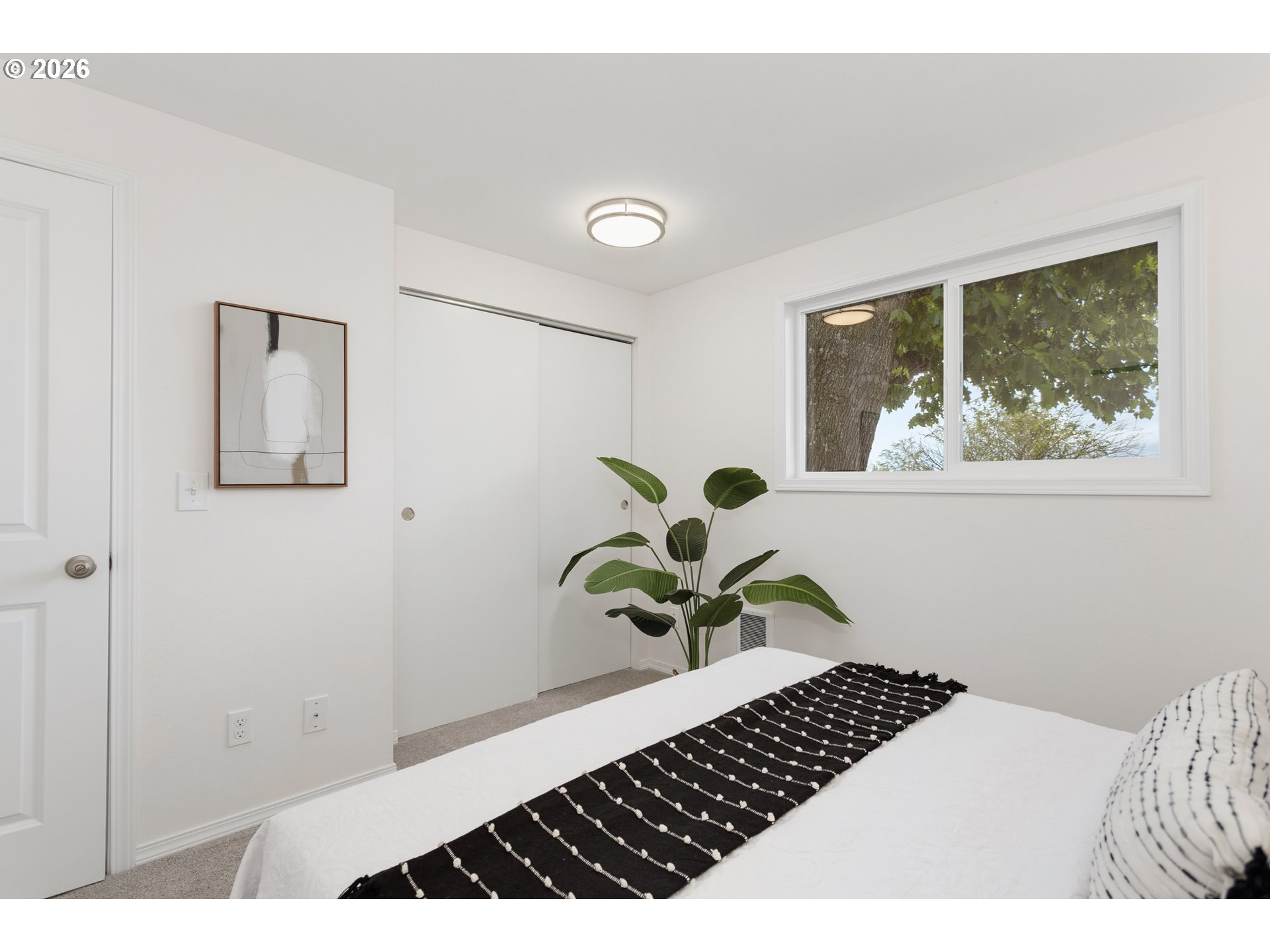 Bedroom with a white bed, black patterned runner, sliding closet doors, and a large window beside a leafy plant.