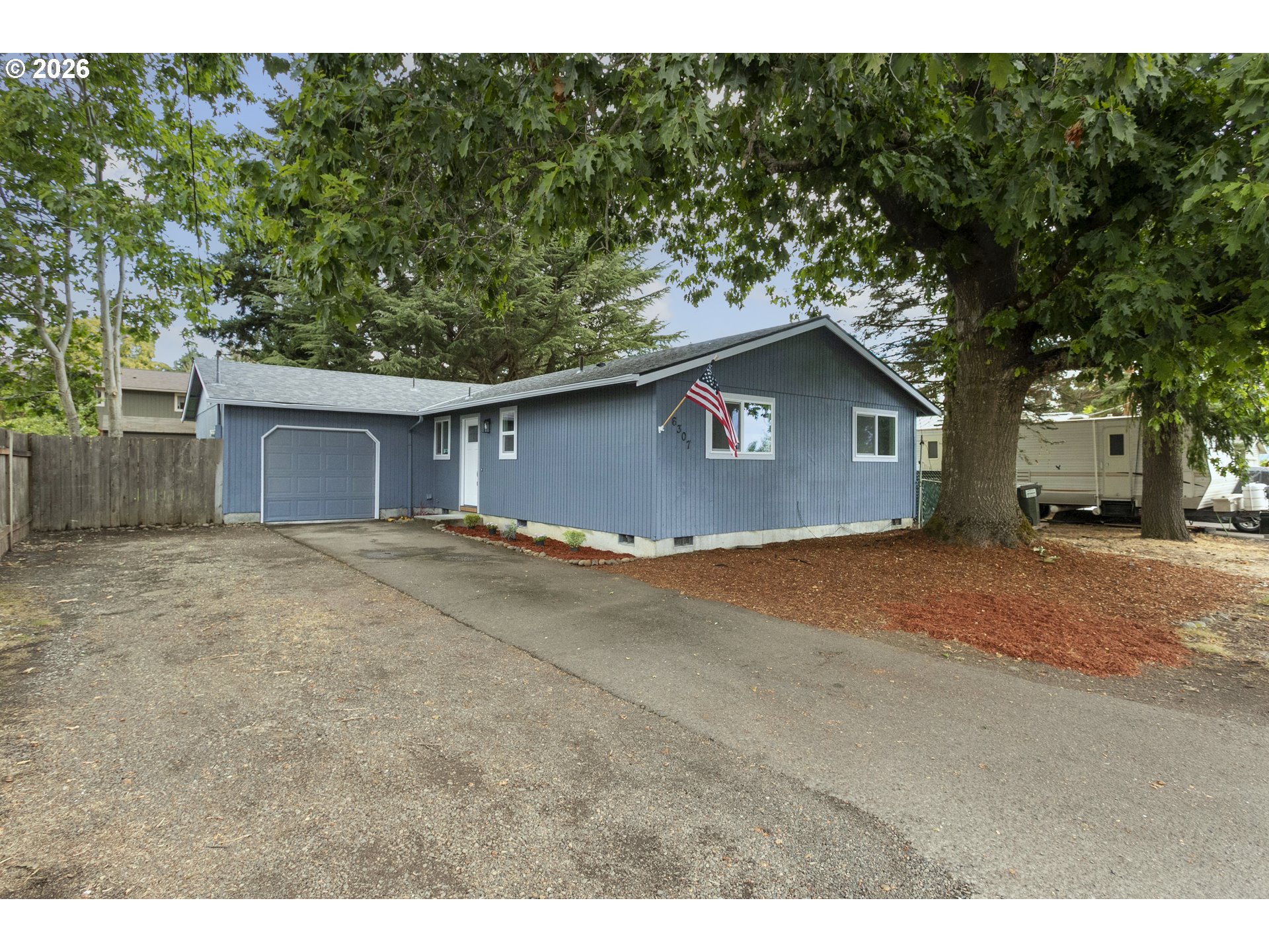 Blue single-story house with an attached garage, American flag, and large shade tree.