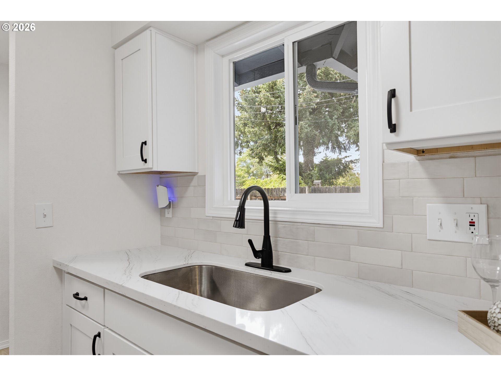 Kitchen sink beneath a large window, with a black faucet, white cabinets, and veined countertop.