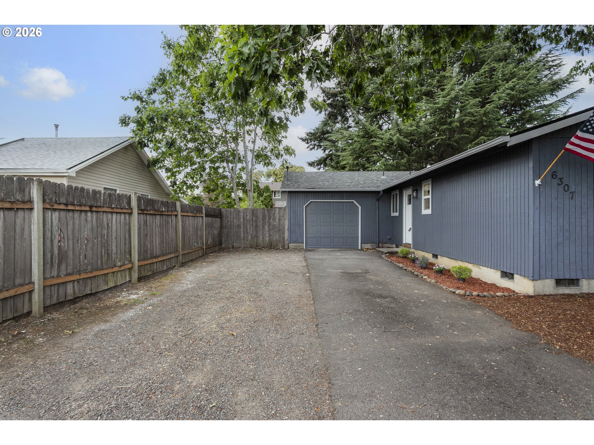 Blue-gray house with attached garage, broad driveway, gravel parking area, and wooden fence.