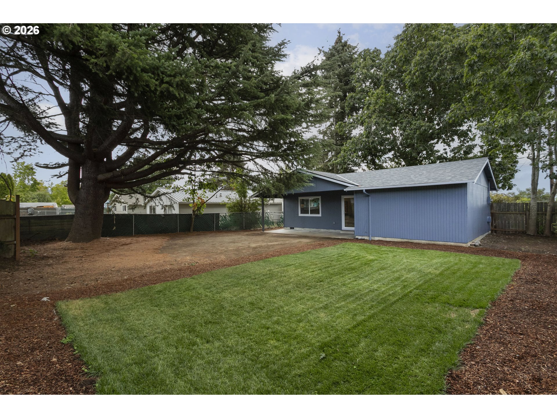 Blue single-story home at the edge of a fenced backyard with a broad lawn and large evergreen tree.