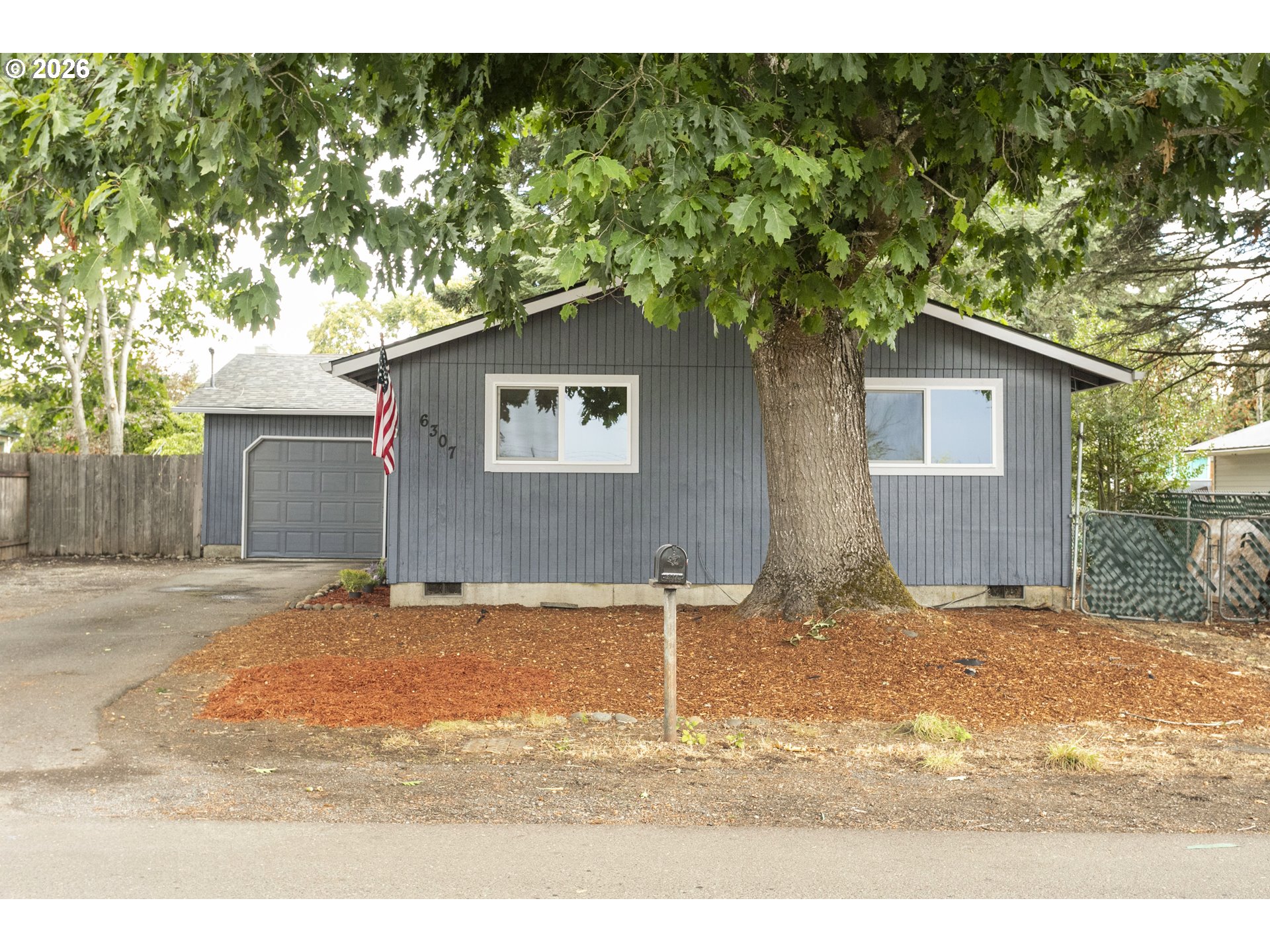 Gray single-story house with a garage and American flag, partly screened by a large mature tree.