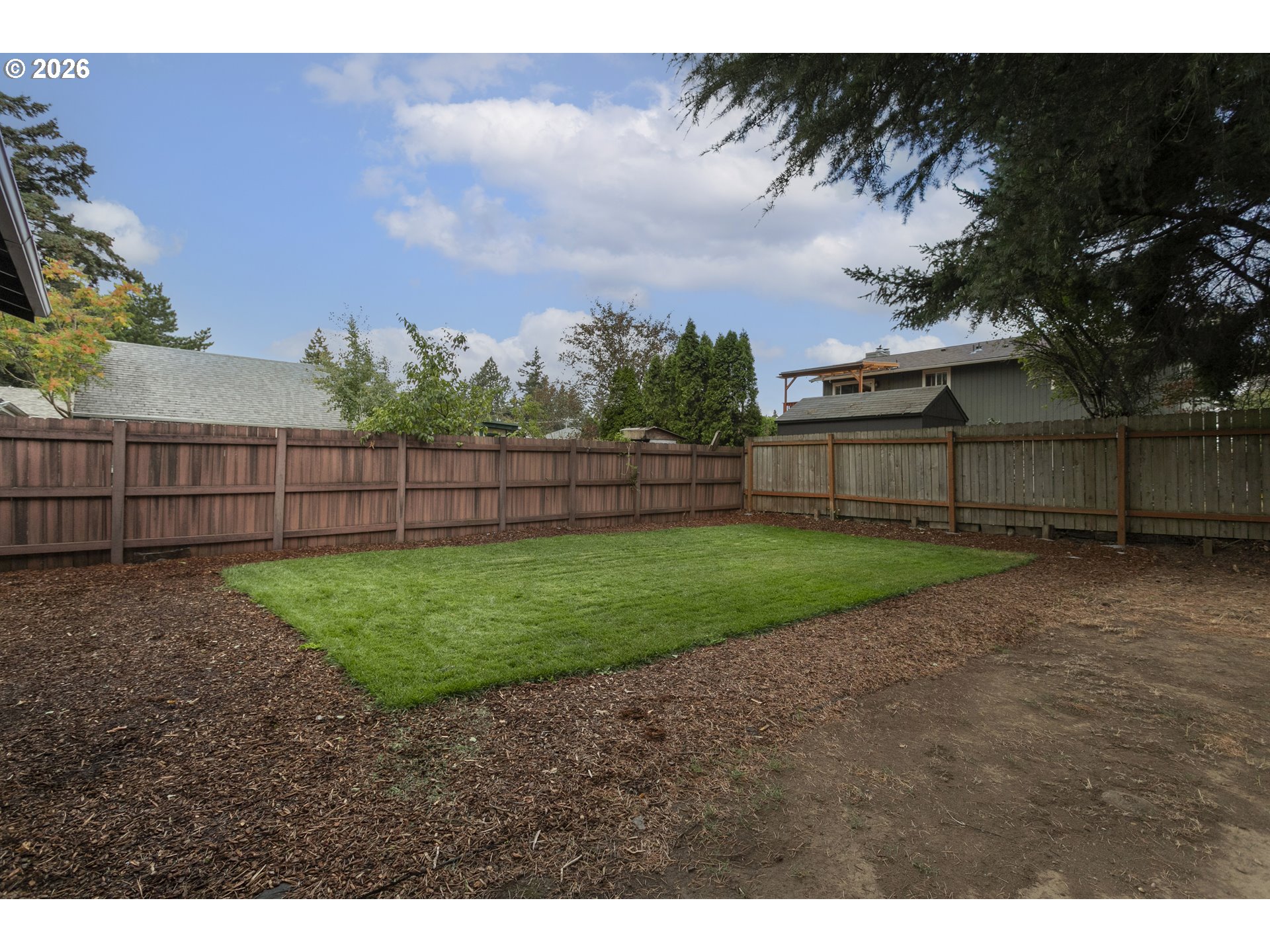 Fenced backyard with a rectangular green lawn, mulched ground, and mature trees.