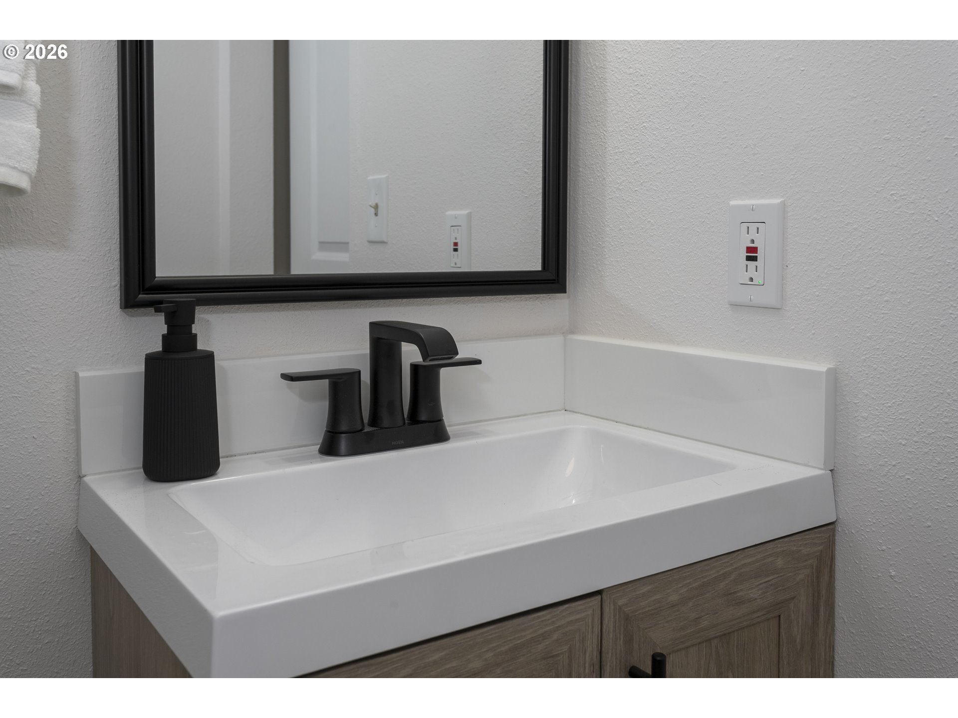 Bathroom vanity with a white sink, black faucet, and wood-grain cabinet beneath a black-framed mirror.