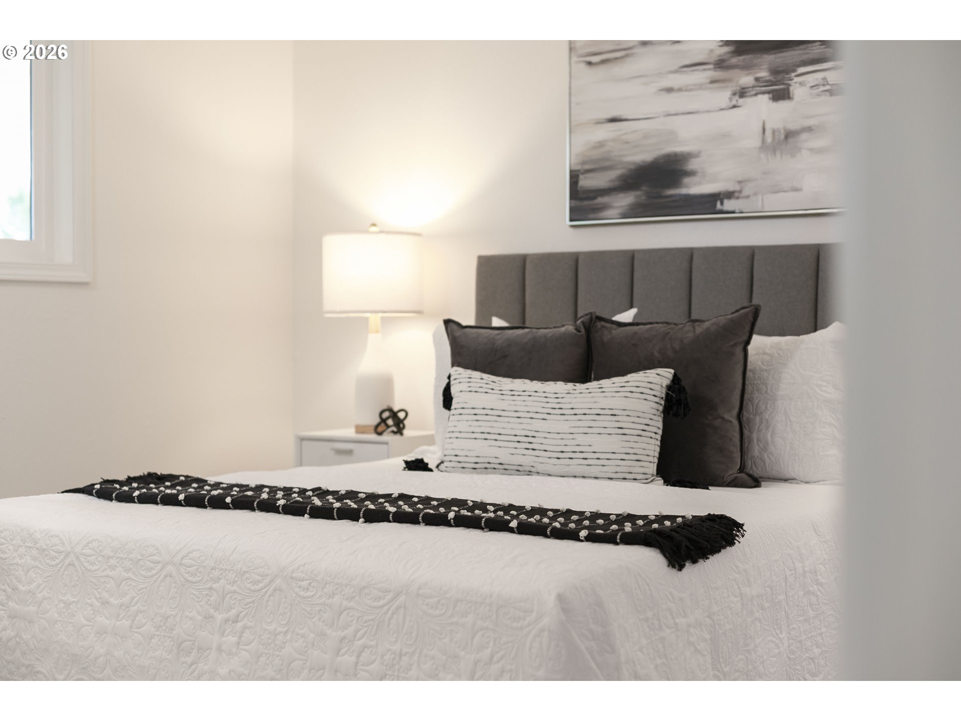 Staged bedroom with a quilted white bed, gray headboard, layered pillows, and bedside lamp.