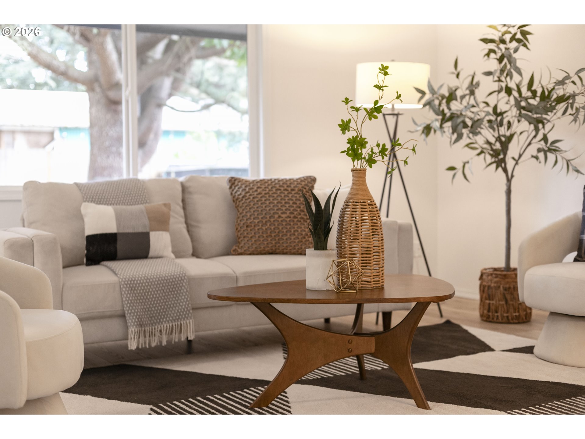 Staged living room with a cream sofa, wooden coffee table, large windows, and leafy plants.