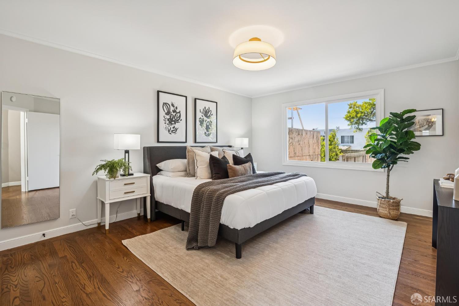 Staged bedroom with a dark upholstered bed, hardwood floors, and a wide window overlooking greenery.