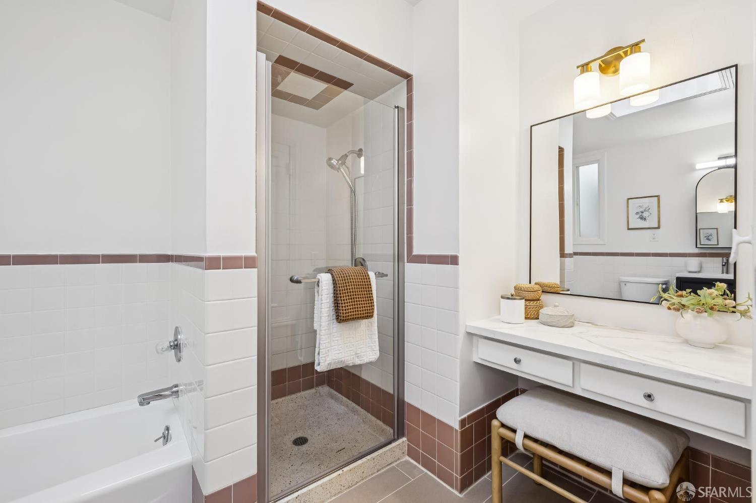 Bathroom with glass shower, bathtub, and white vanity accented by mauve-brown tile and brass lighting.