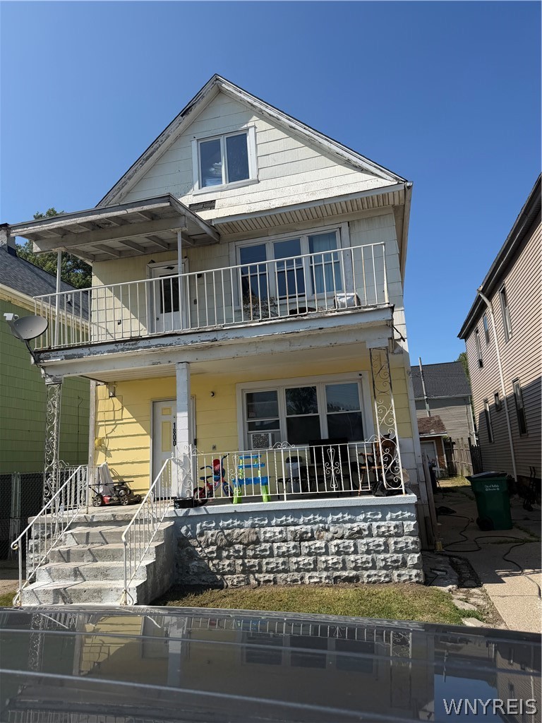 Two-story pale yellow house with a stone foundation, front porch, and second-floor balcony.