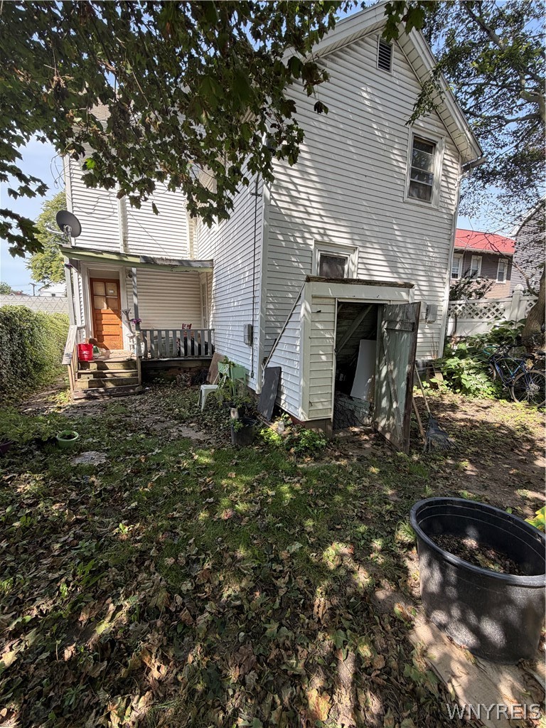 Rear of a white-sided house with a wooden porch and open basement entrance, seen from a leaf-covered yard.