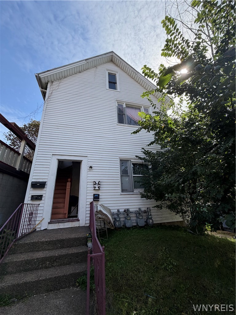 White-sided multi-story house with an open ground-floor entrance, concrete steps, and a leafy tree beside it.