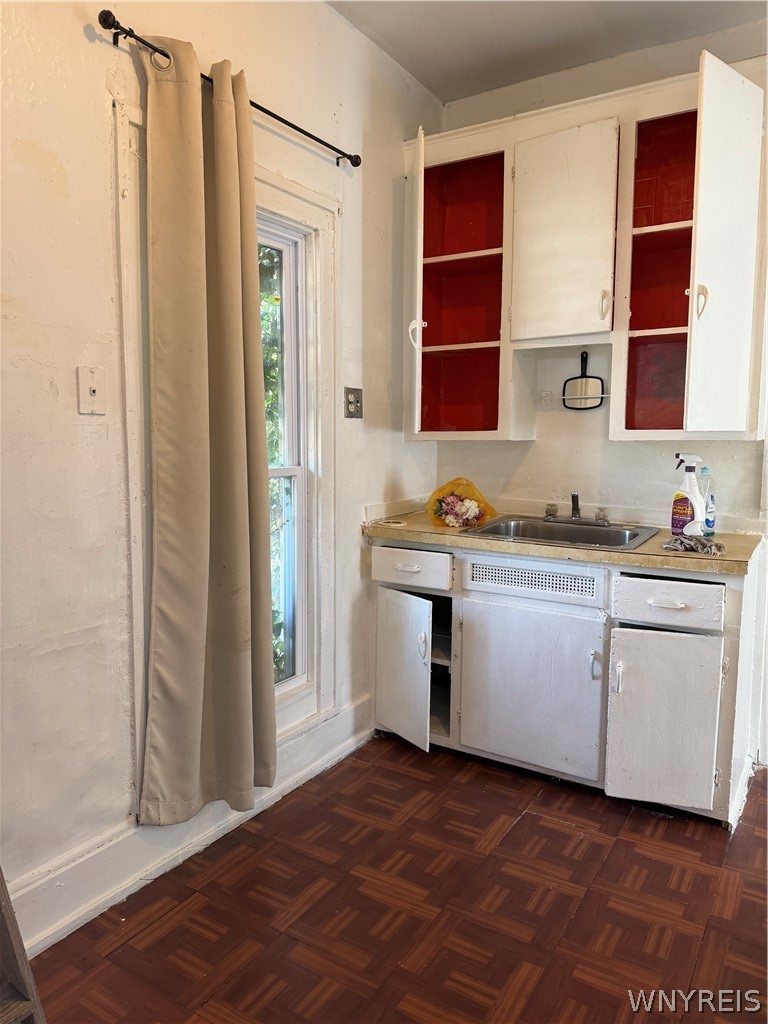 Small kitchen with open white cabinets, a stainless-steel sink, curtained window, and dark parquet floor.